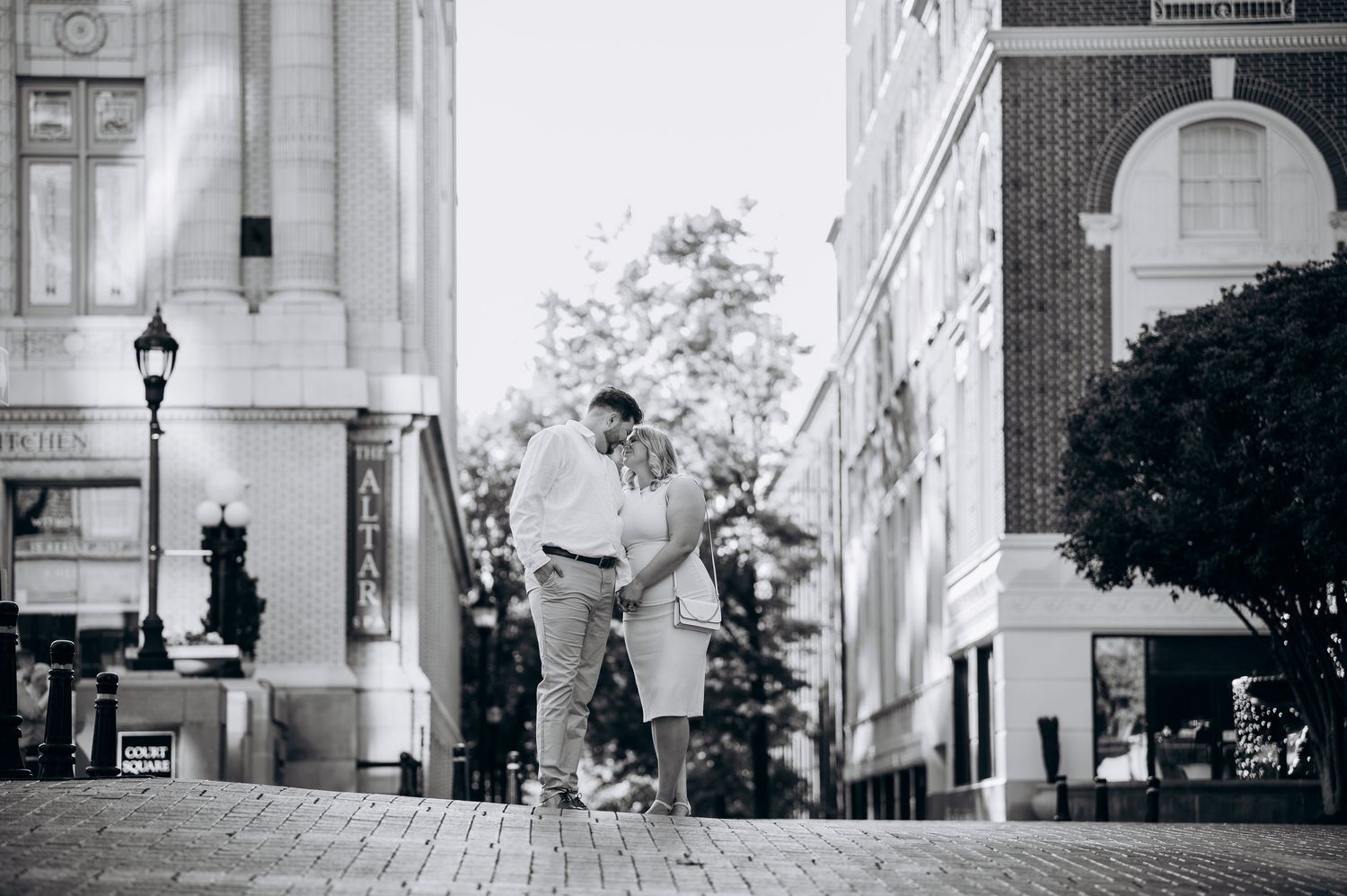 Black and white romantic photo of a couple embracing on a cobblestone street between elegant buildings.