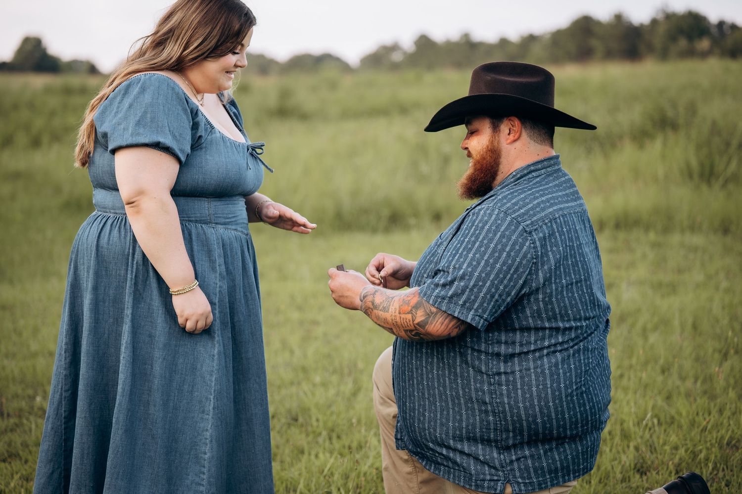 Man kneeling on one knee presenting an engagement ring to woman in a green meadow.