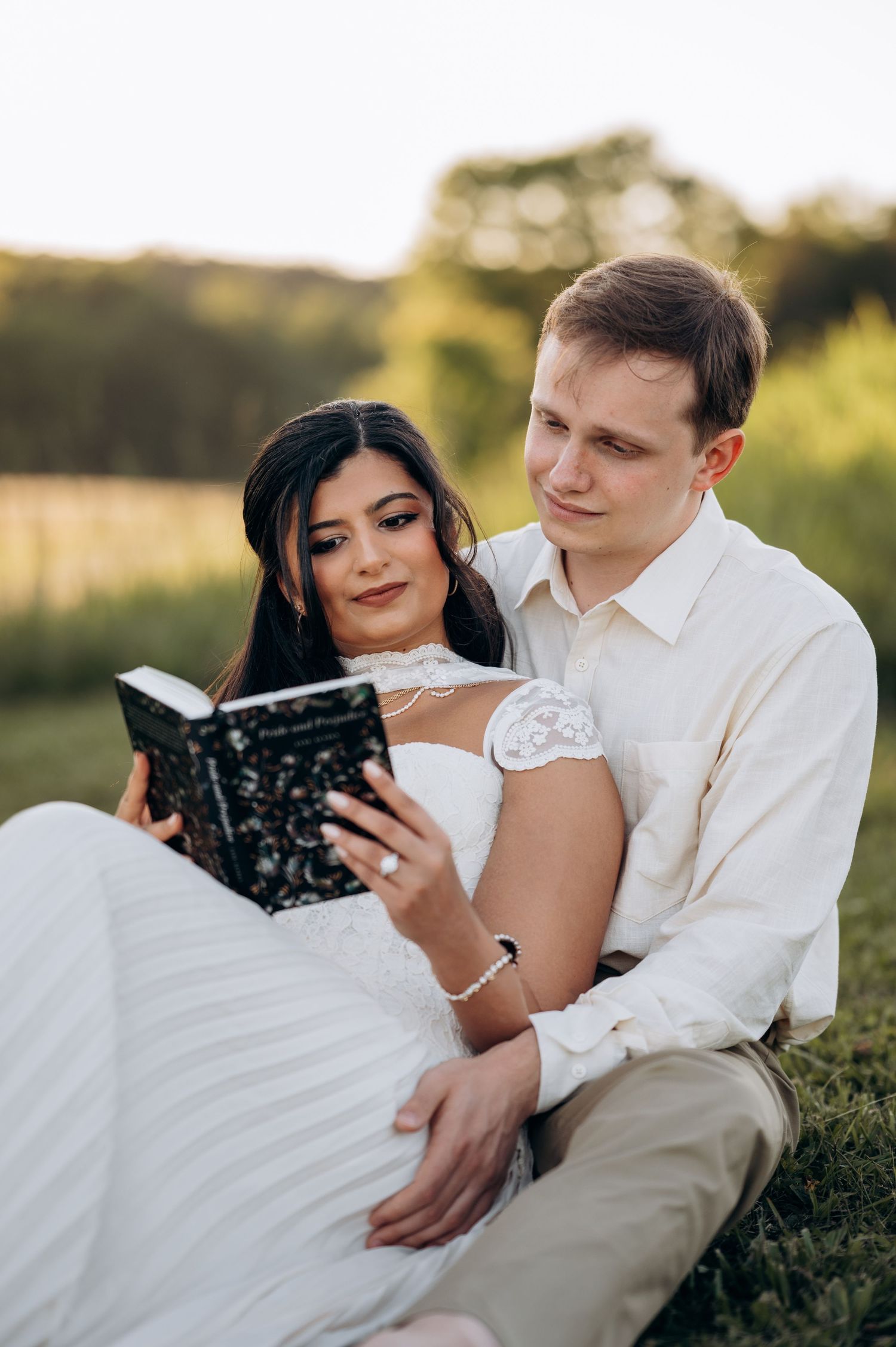 Couple in wedding attire sitting on grass reading Pride and Prejudice book together at golden hour.