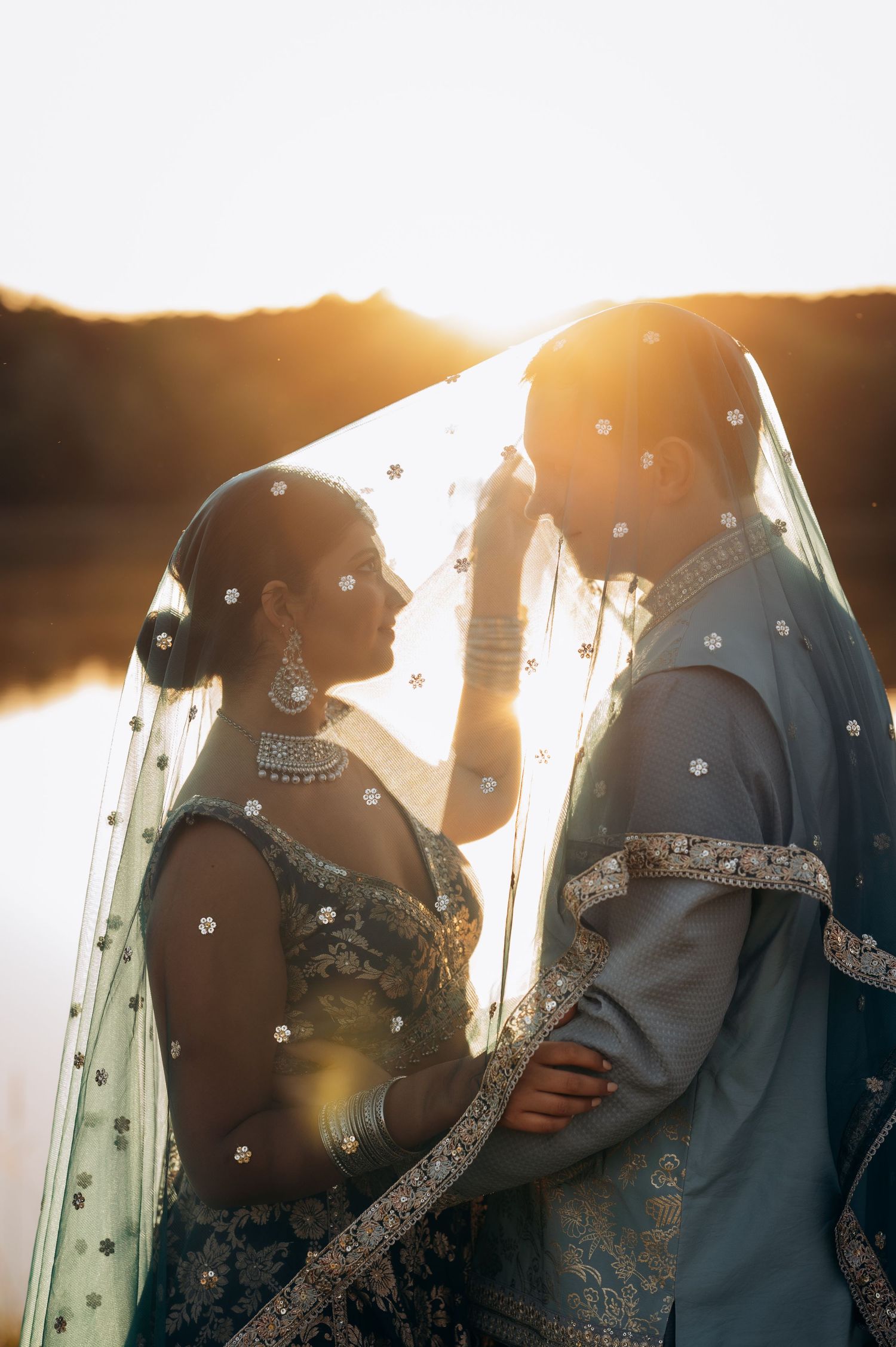 South Asian bride and groom in blue traditional attire share romantic moment under embellished veil at golden sunset.