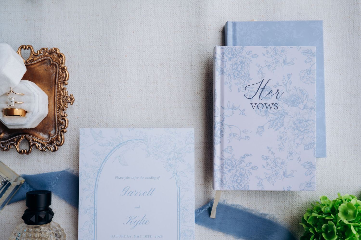 Elegant light blue wedding vow books and invitation suite with floral details arranged on a soft linen background.