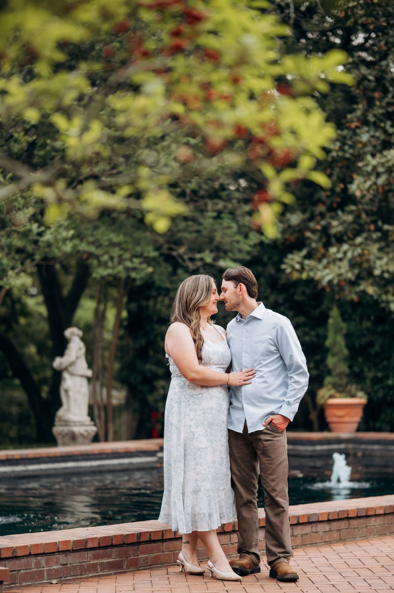 Romantic couple embracing near a fountain with a classical statue in a beautiful garden setting.