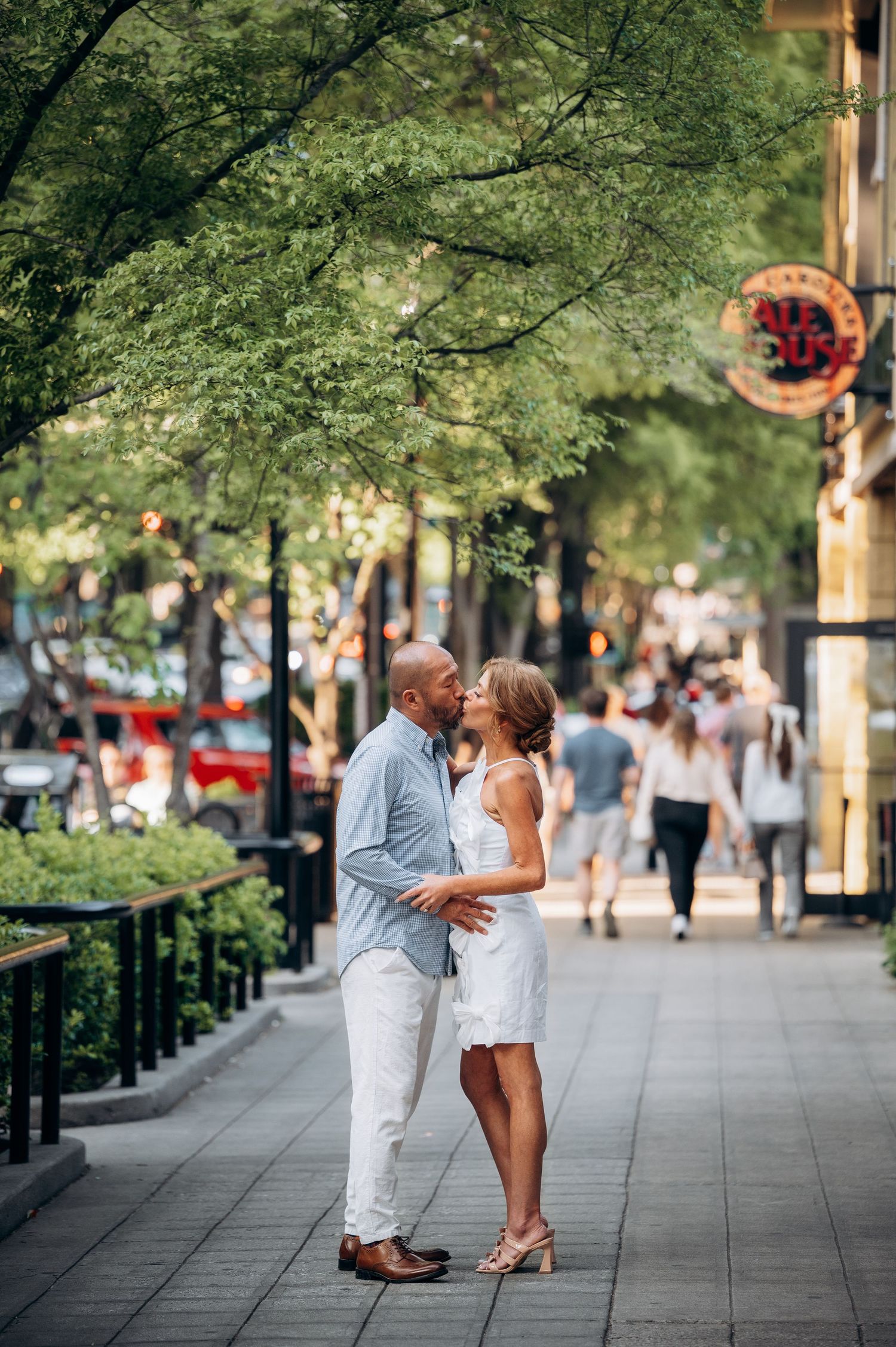 Couple sharing a romantic kiss on a tree-lined urban sidewalk, man in blue shirt and woman in white dress.