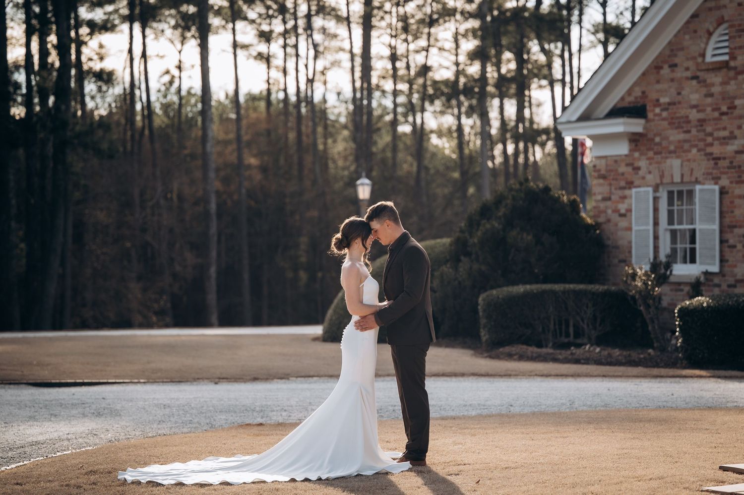 Bride and groom sharing romantic kiss outdoors near brick estate surrounded by tall pine trees.