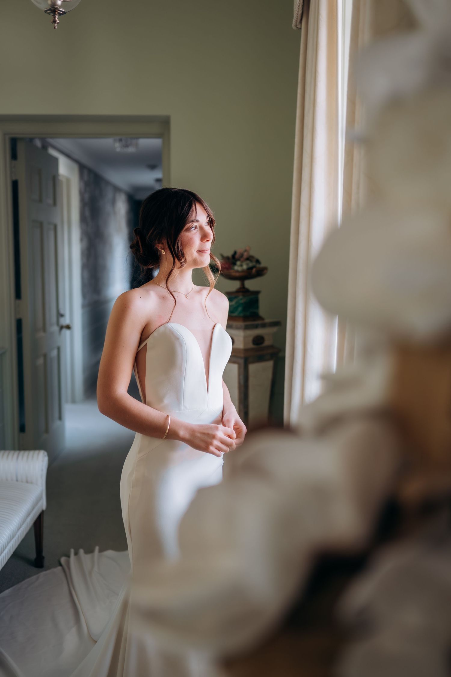 Bride in elegant strapless white gown gazing out window in soft natural light before wedding ceremony.