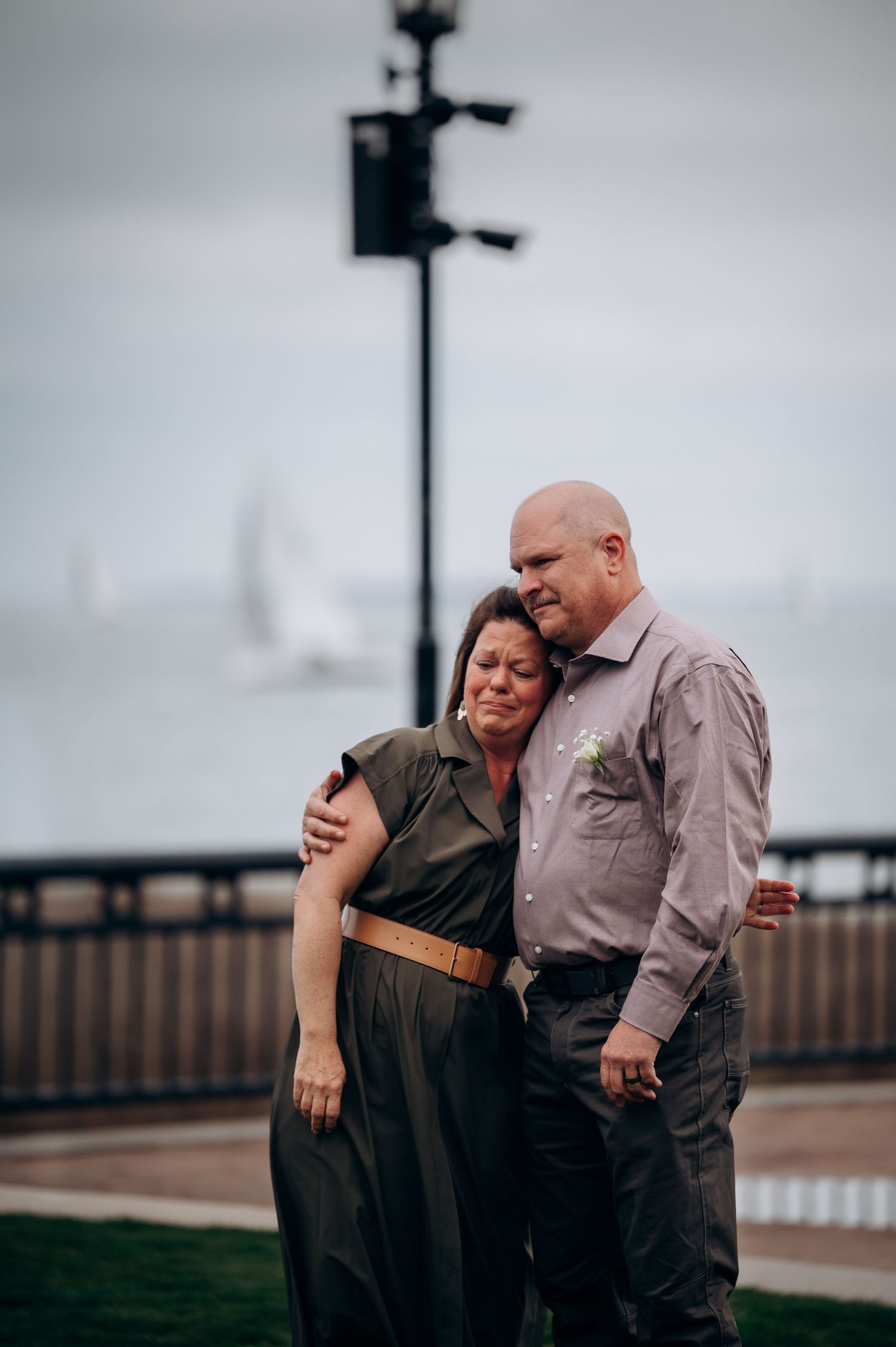 Couple embracing outdoors near waterfront, woman in olive jumpsuit leans on mans shoulder near harbor.