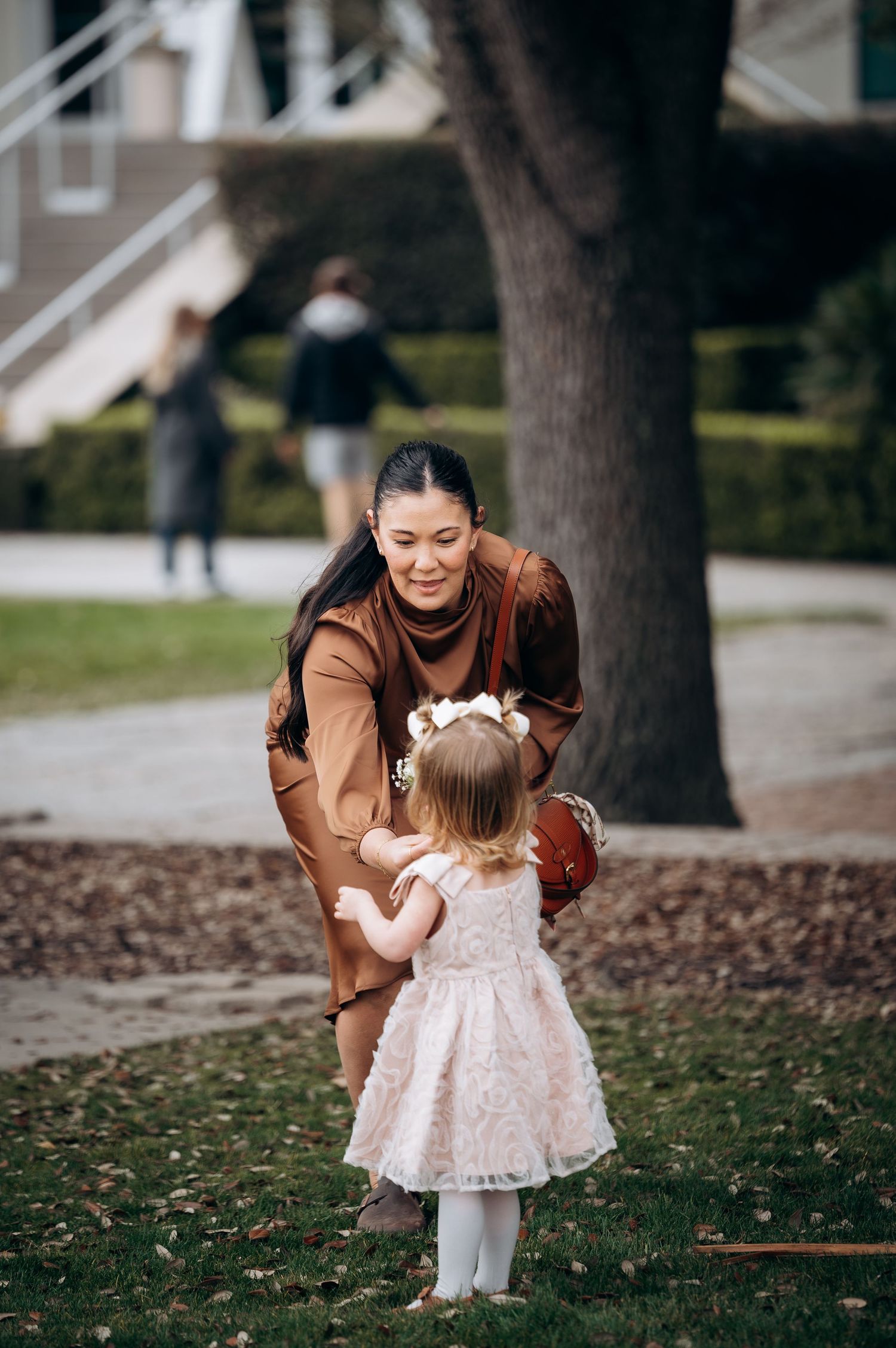 Woman in brown dress bends down to greet a toddler girl in white lace dress with flower crown in a park.