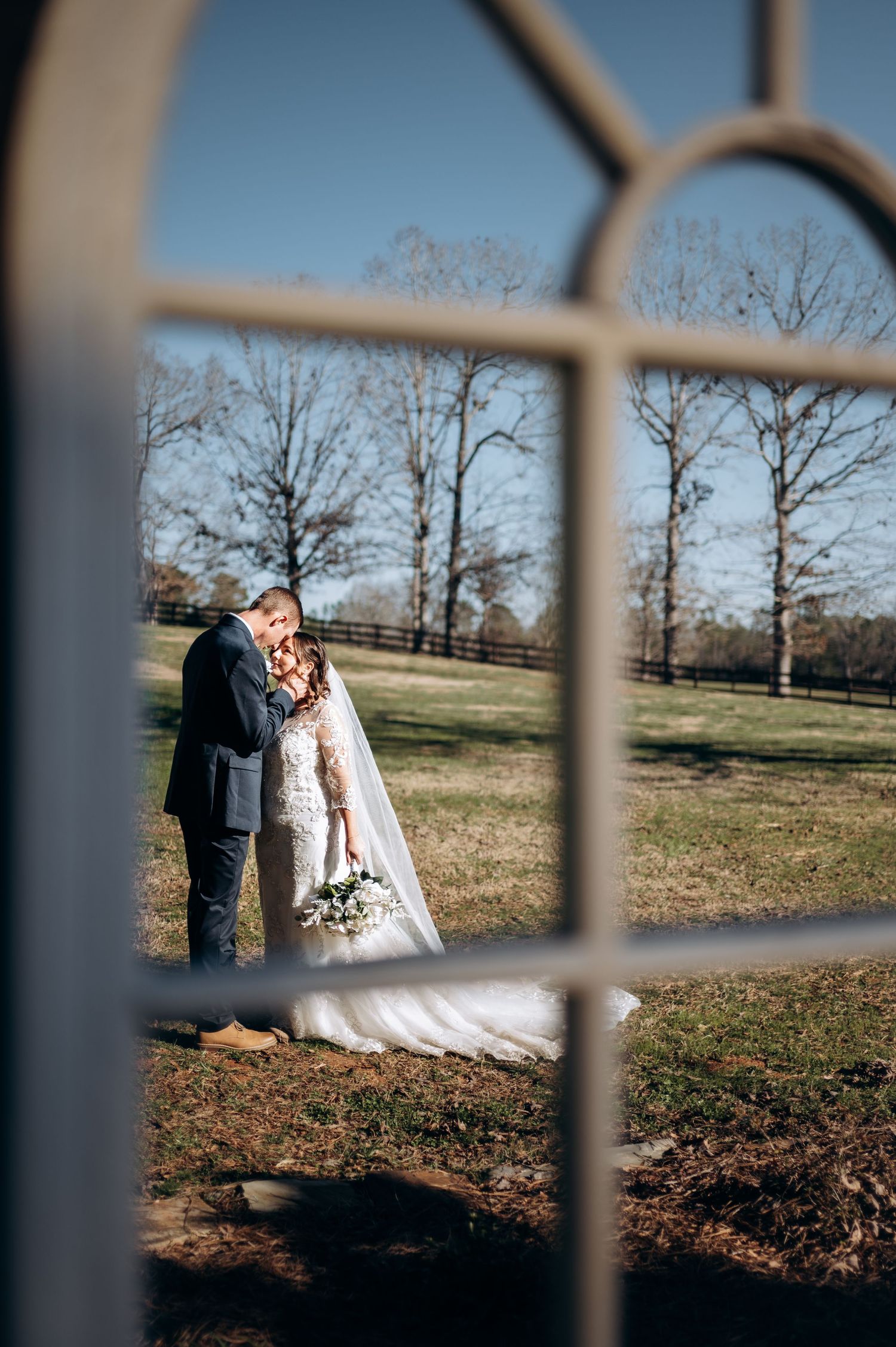 Bride and groom share a romantic kiss framed through a vintage window at an outdoor rustic wedding venue.