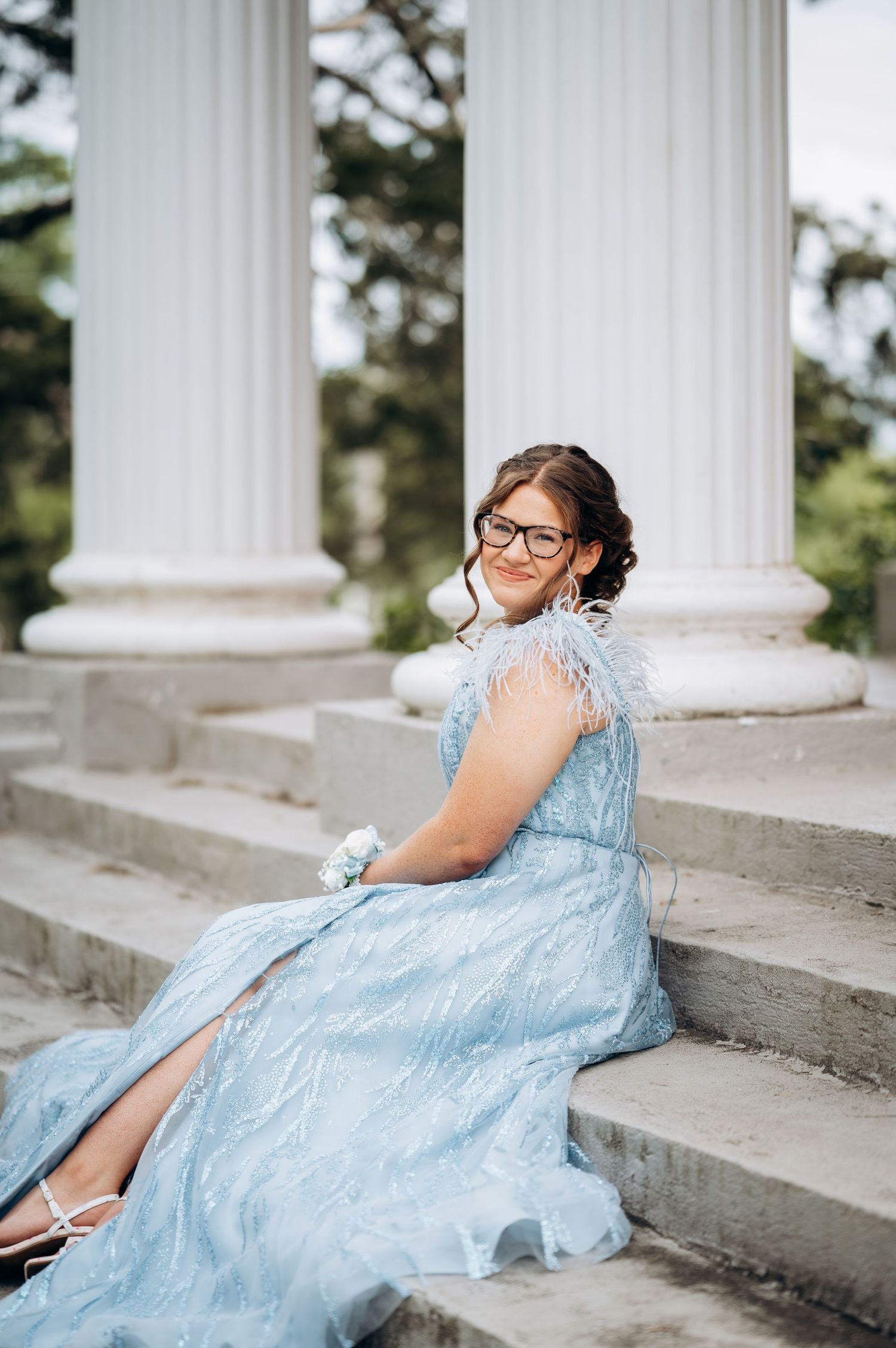 Young woman in a sparkling light blue prom dress with feather details sits gracefully on stone steps by white columns.