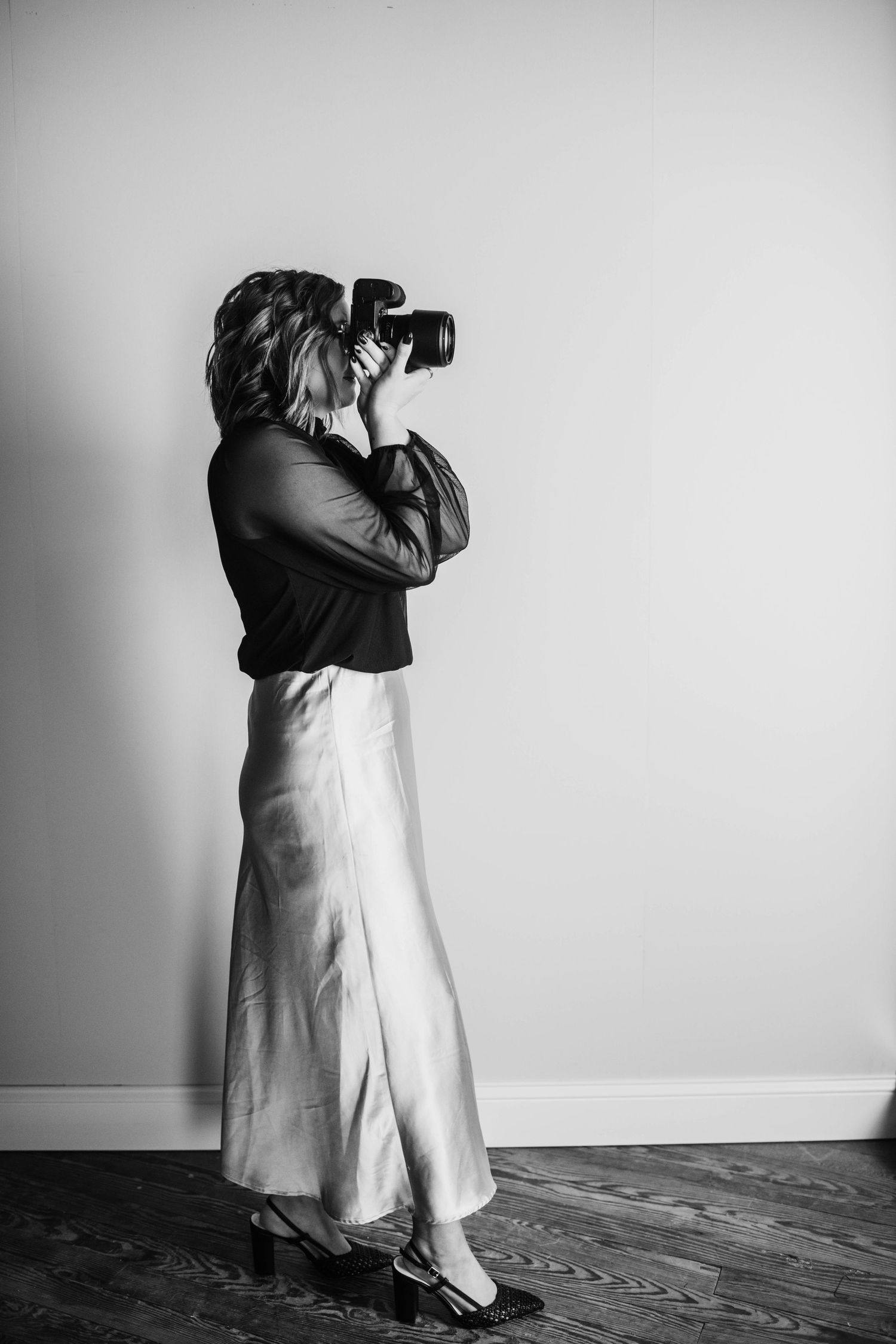 Silhouette of a photographer in a flowing skirt taking pictures against a white wall.
