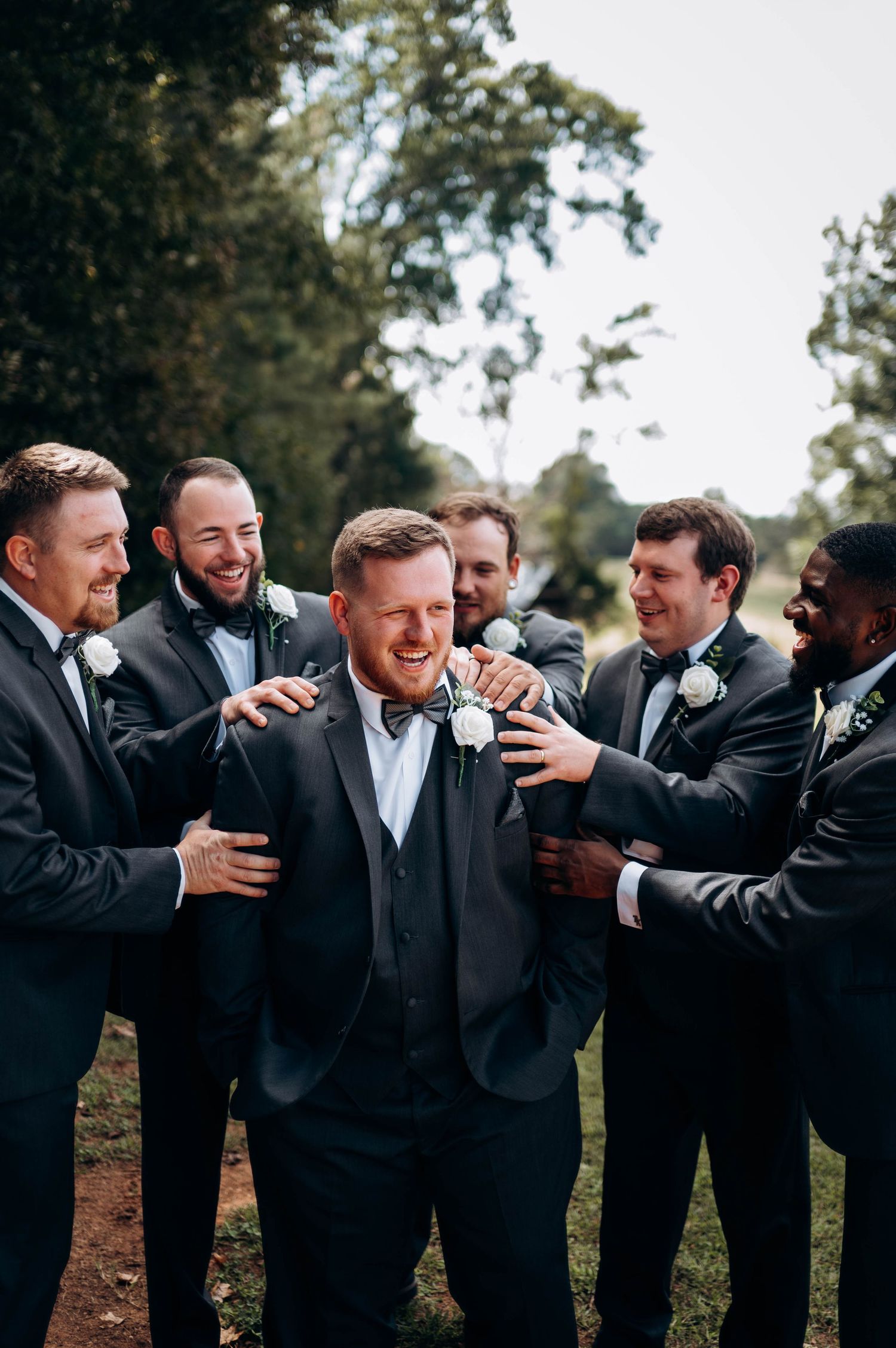 Groomsmen in black tuxedos laugh together during outdoor wedding photo session.