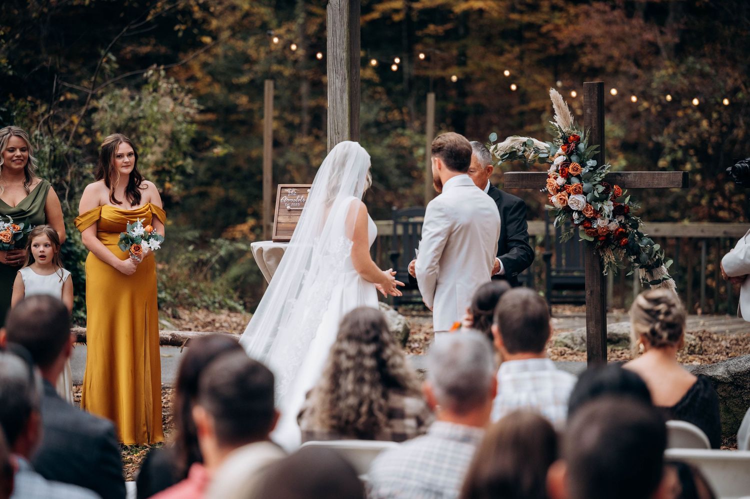 Outdoor autumn wedding ceremony with guests seated under string lights.
