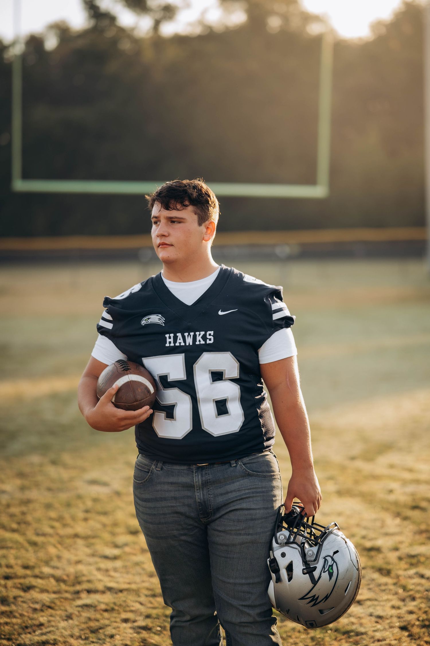 Football player in black Hawks uniform stands on grass field holding helmet and ball.
