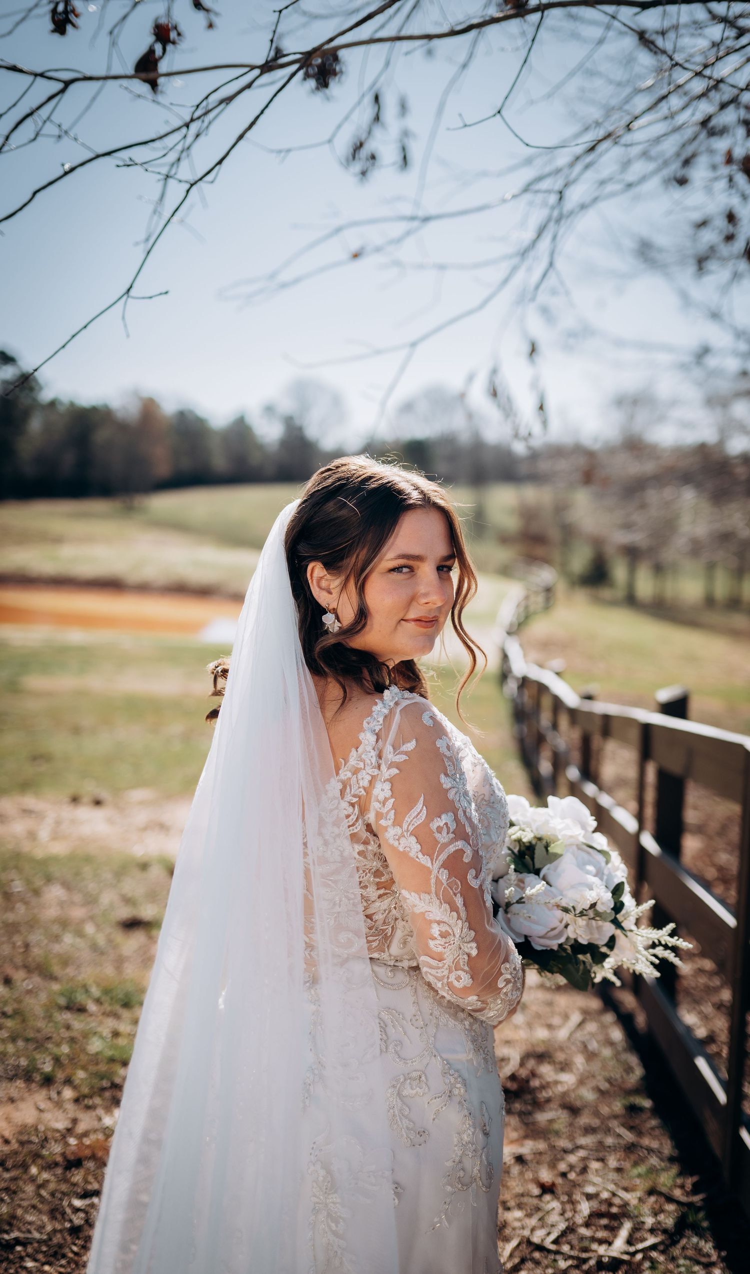 Ethereal bridal portrait in lace dress with veil against autumn landscape and wooden fence.