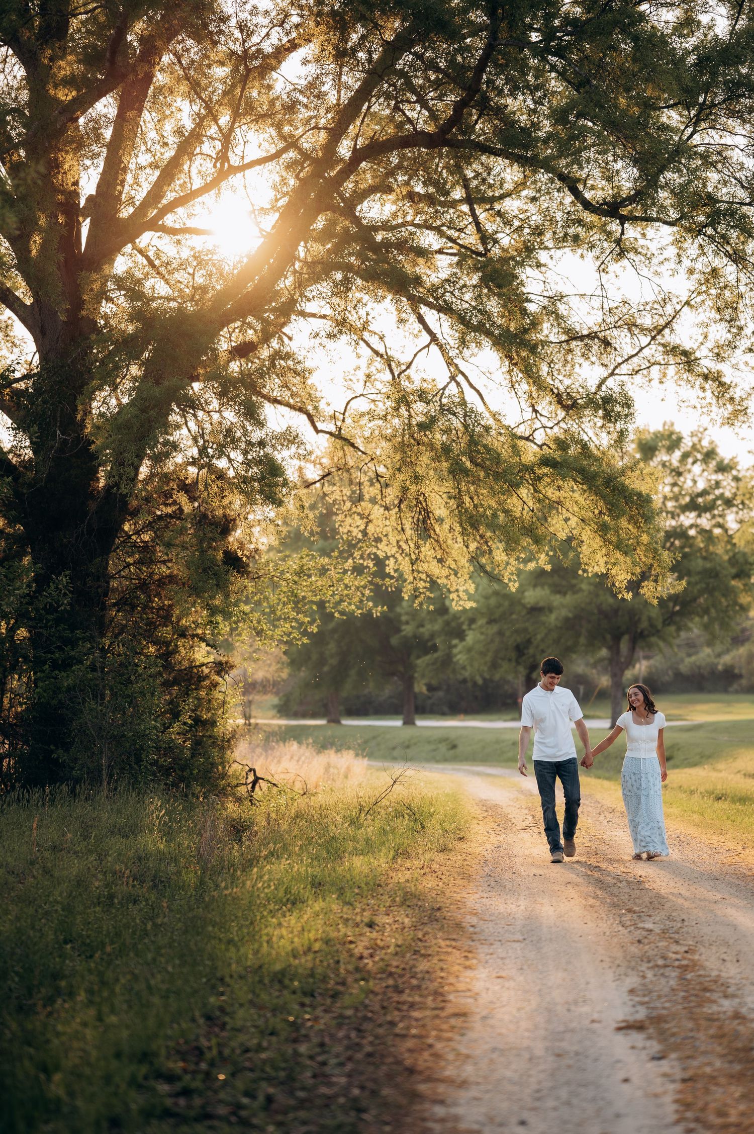 Couple holding hands walking down a sunlit dirt path surrounded by lush green trees during golden hour.