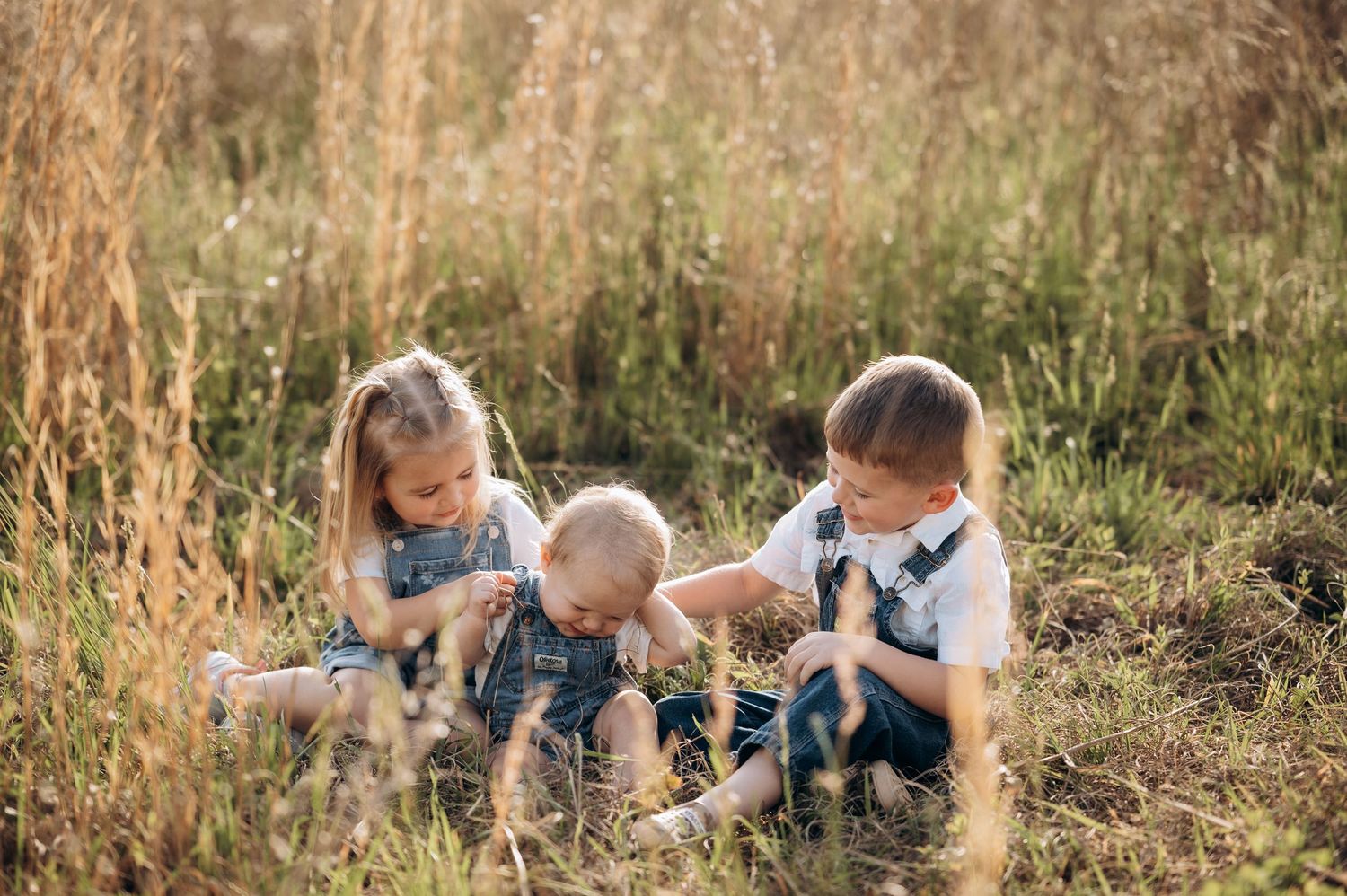 Three young children sit in a sunlit meadow, playing together in matching denim overalls.