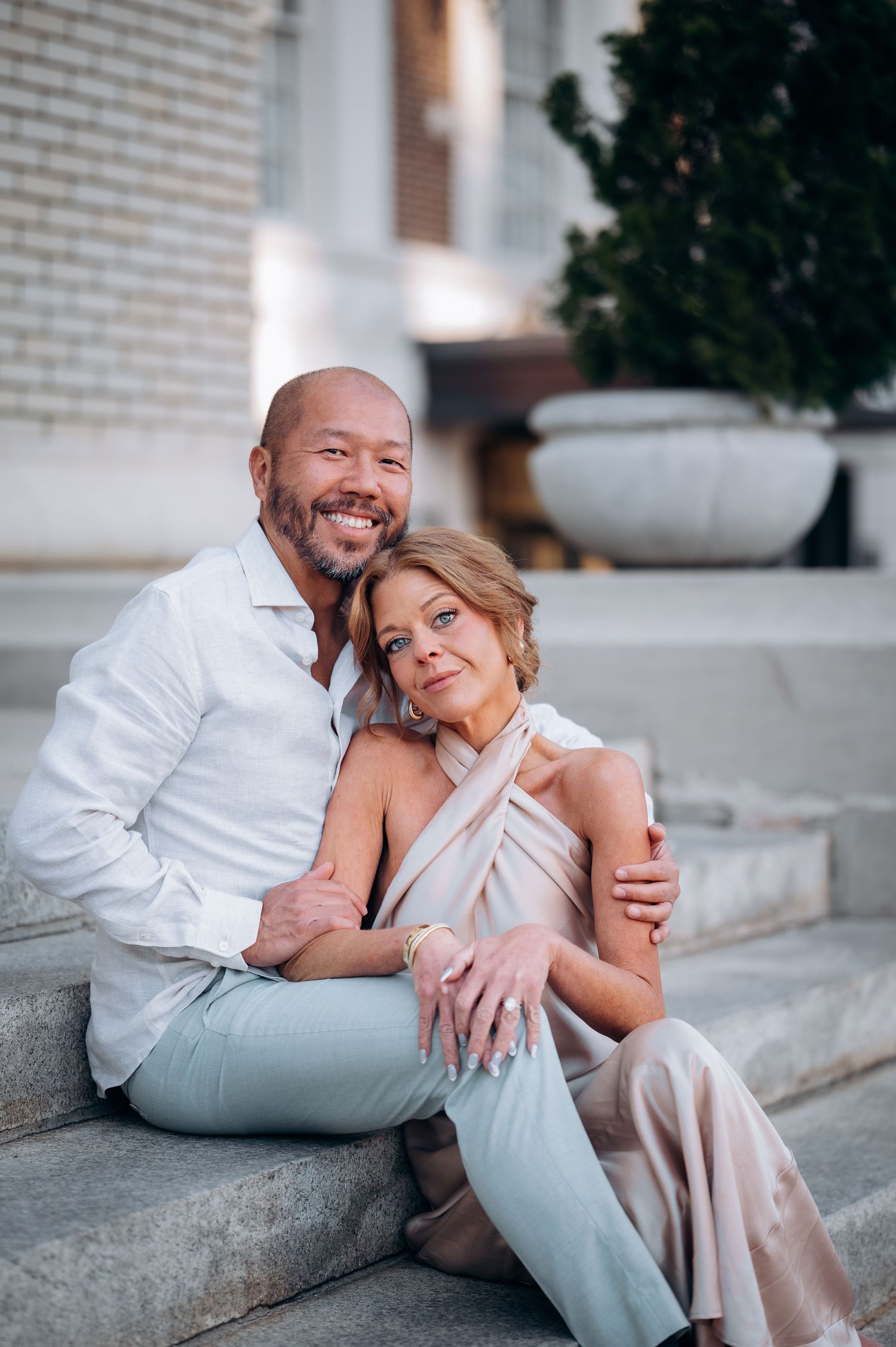 A happy couple sits on stone steps outdoors, the man embracing the woman in elegant attire.