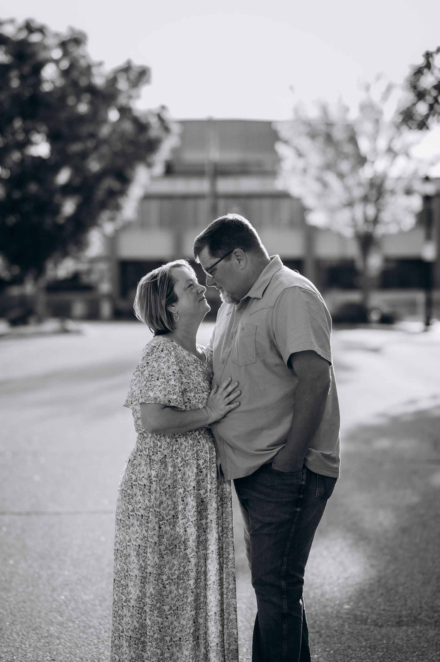 Black and white photo of a couple sharing a romantic moment on a street, woman in floral dress, man in casual shirt.