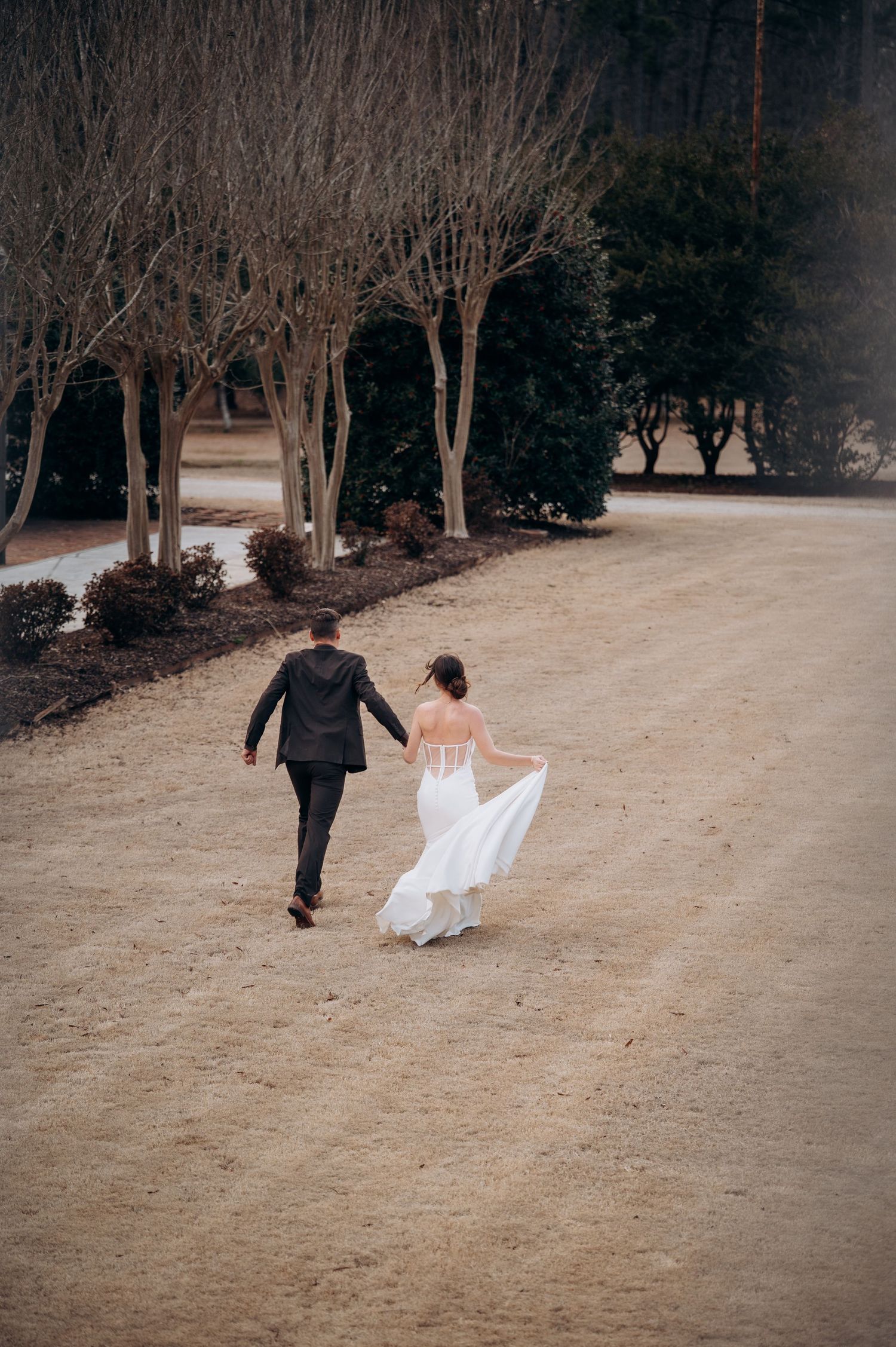 Bride and groom walking hand in hand down a gravel path surrounded by bare trees on their wedding day.