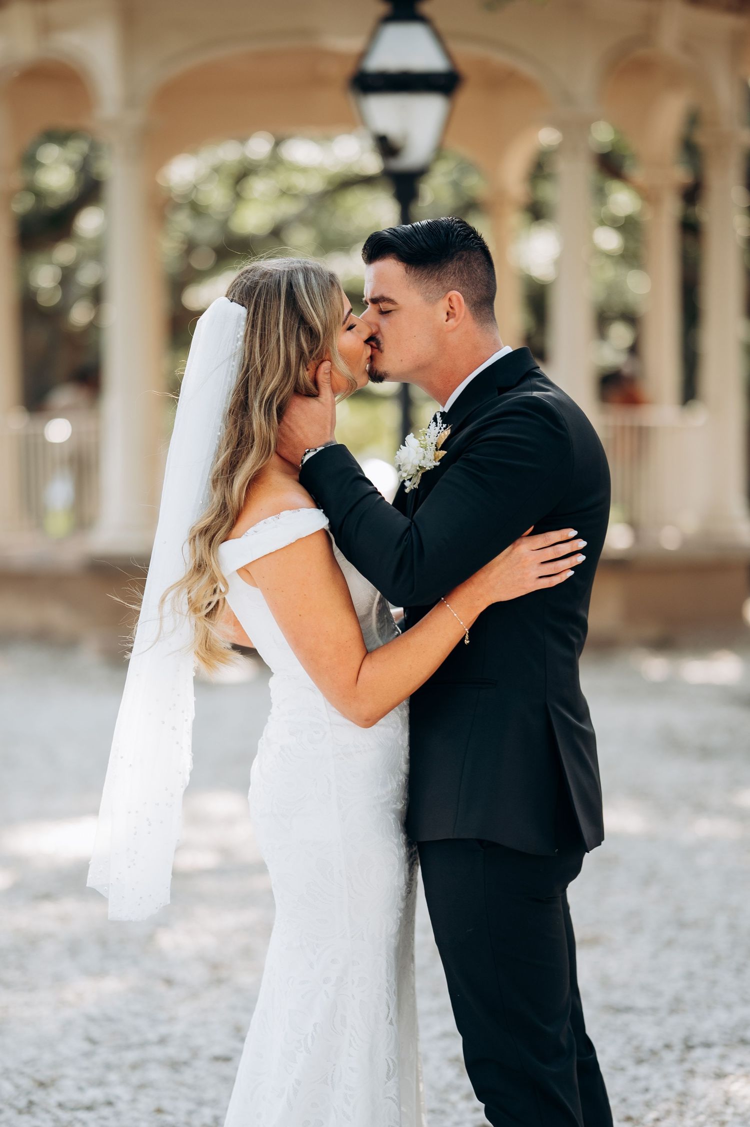 Bride and groom share a romantic kiss outdoors, bride in white lace gown with veil, groom in black suit with boutonniere.