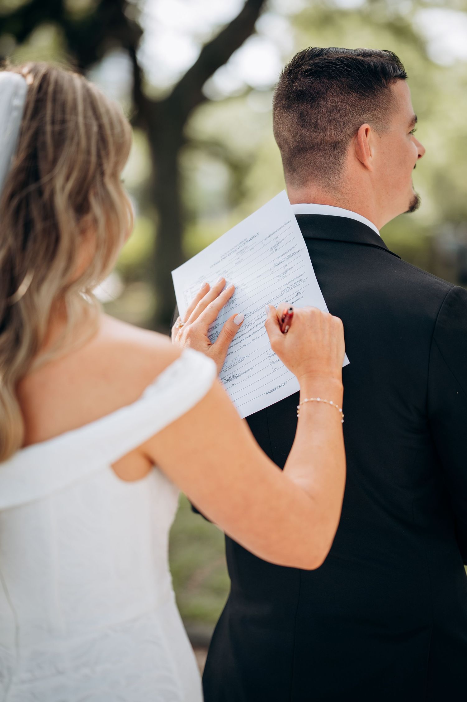 Bride signs wedding certificate on groom's back during outdoor ceremony under lush green trees.