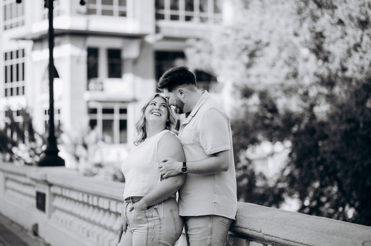 Black and white photo of a couple sharing a tender moment on a bridge, smiling and embracing in an urban setting.