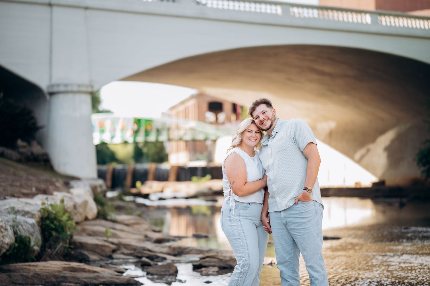 Couple posing together near a creek under a bridge during golden hour engagement photo session.