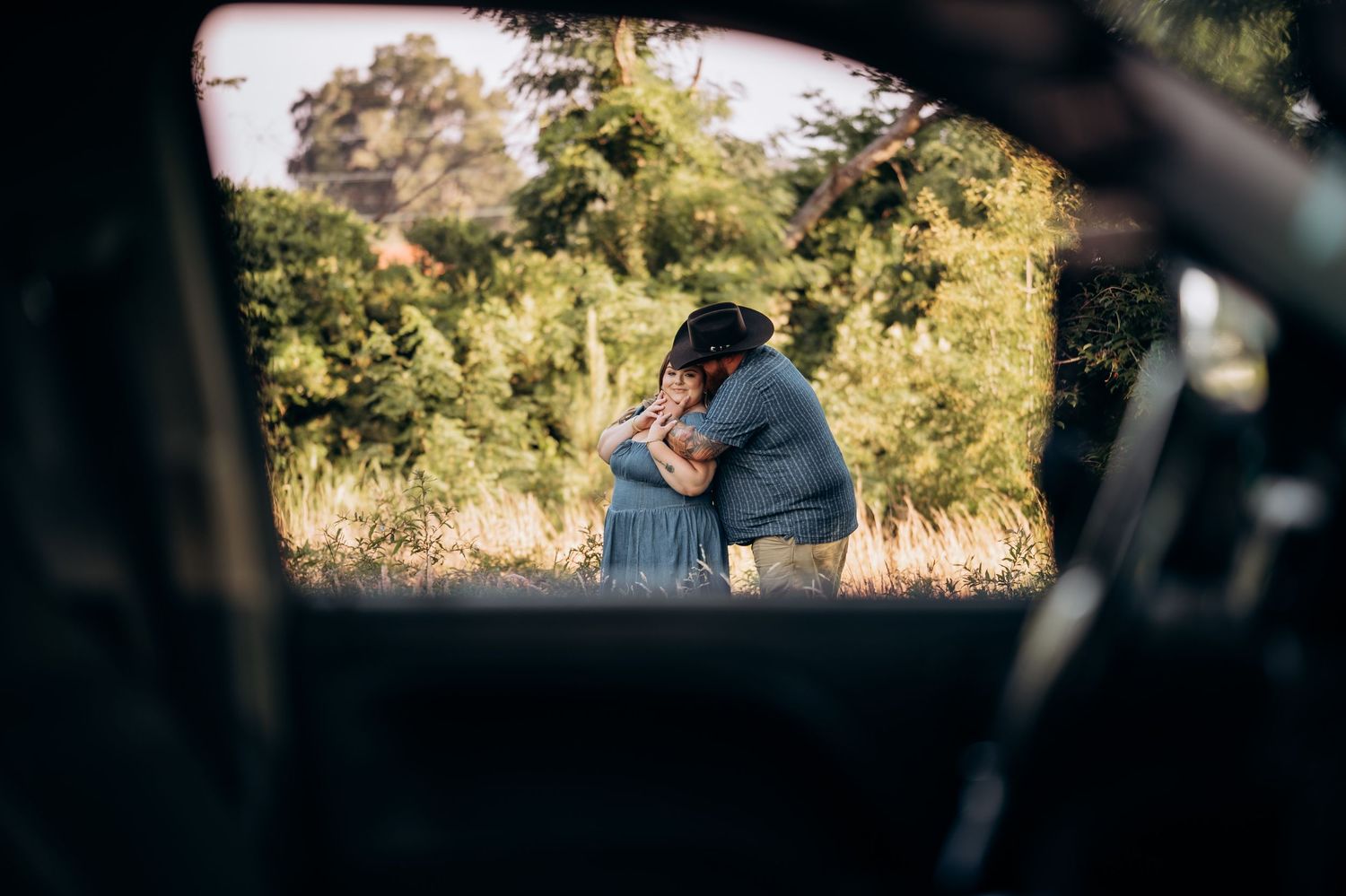 Couple embracing outdoors near a car window surrounded by lush greenery.