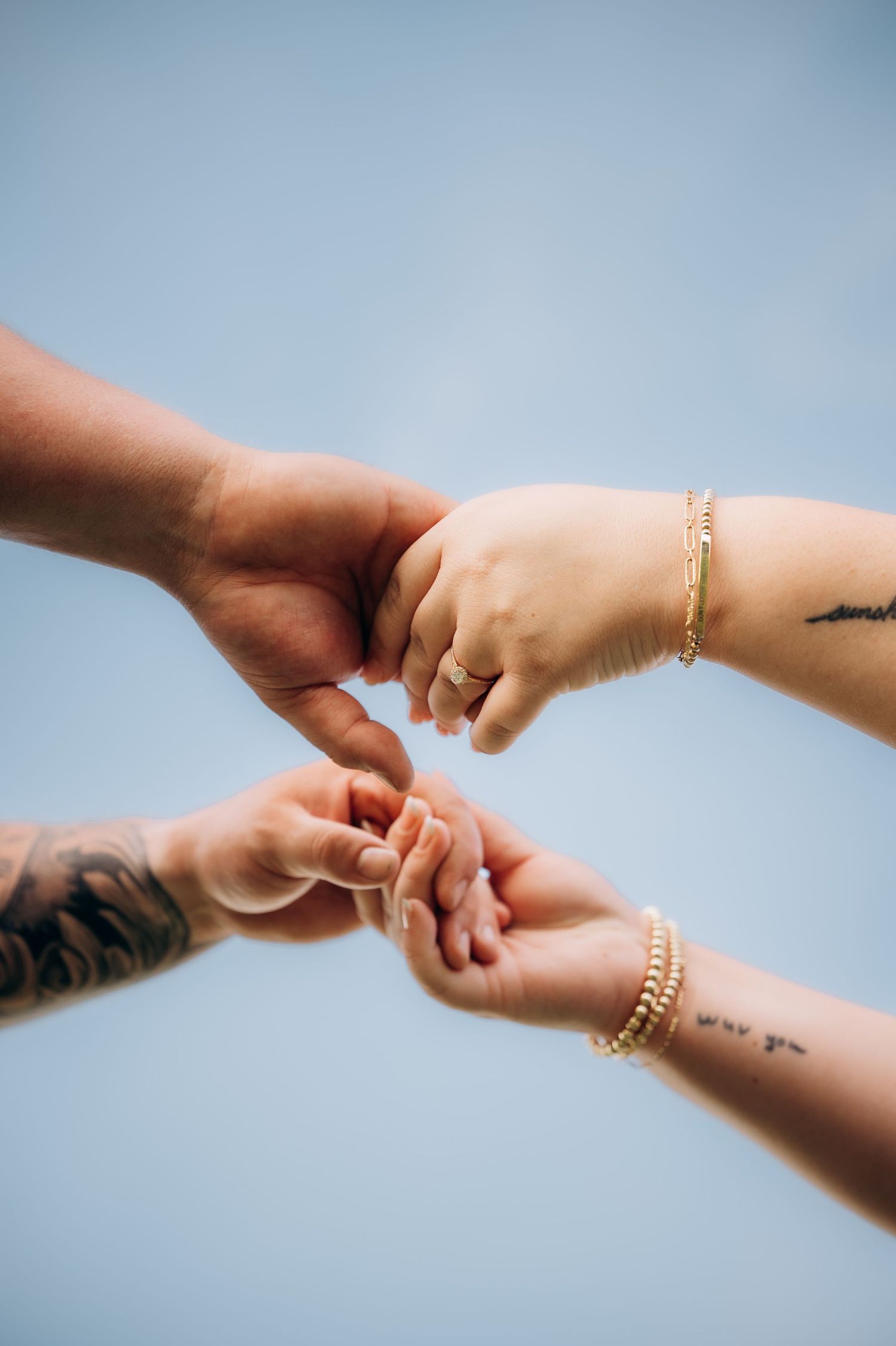 Four hands with tattoos and gold bracelets connecting fingers against a blue sky.