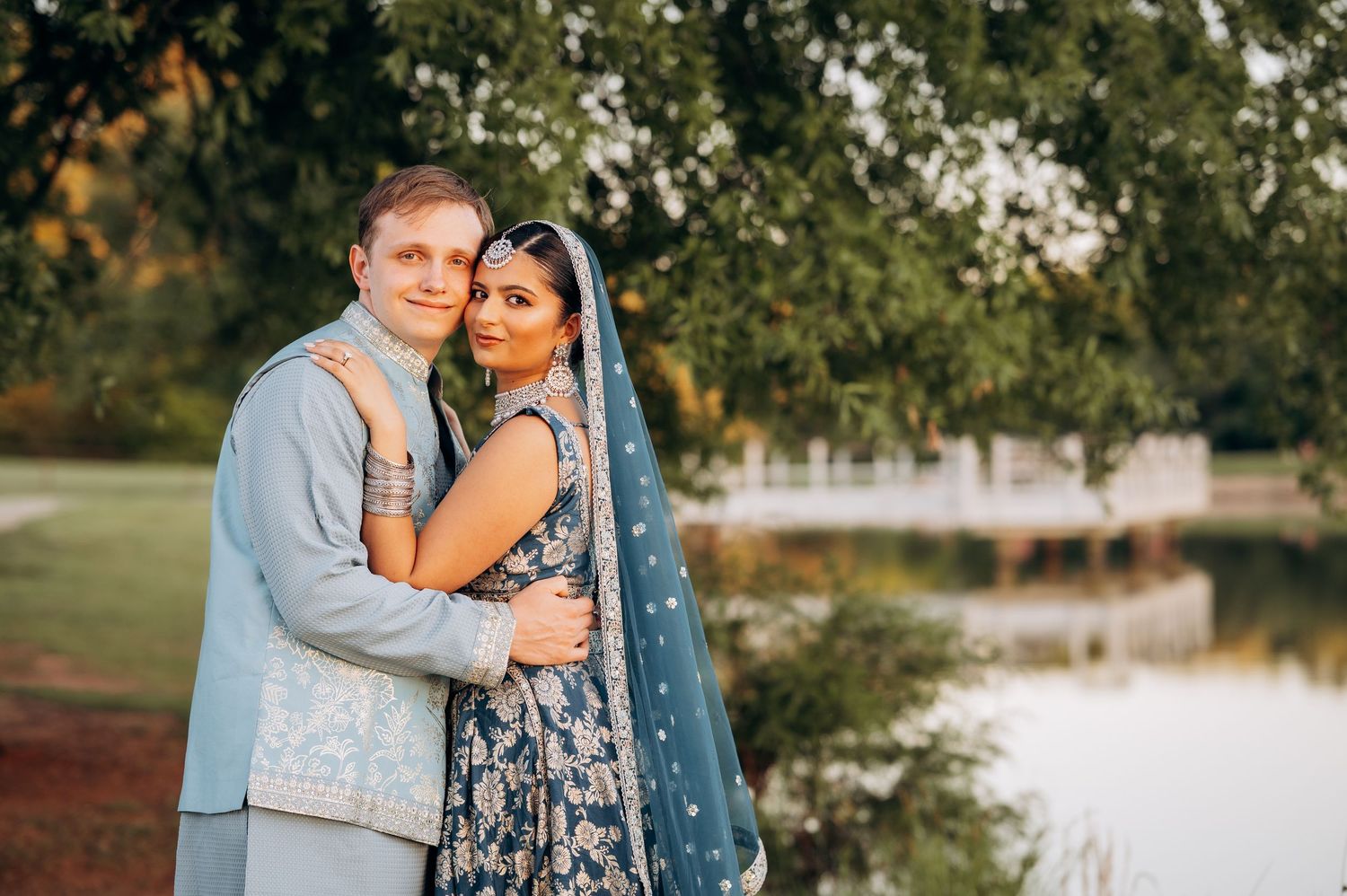 Couple in traditional South Asian wedding attire poses by a lake, woman in blue lehenga with dupatta, man in gray sherwani.