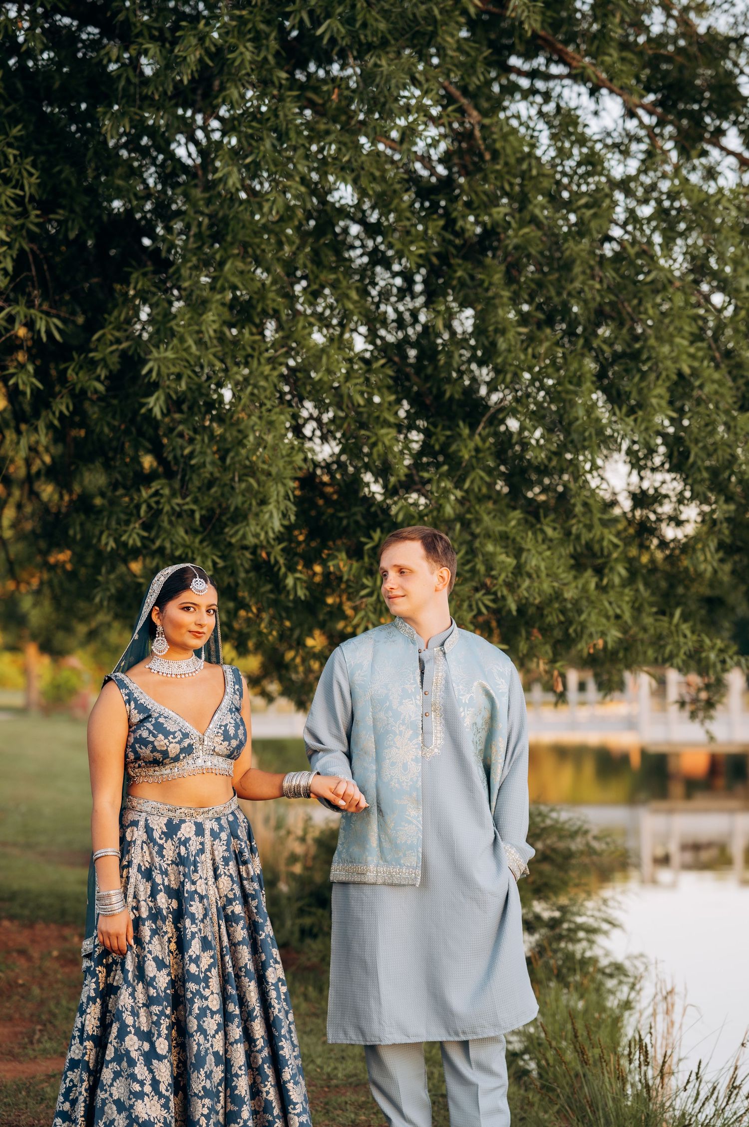 Couple in traditional Indian wedding attire, bride in blue lehenga and groom in kurta, posing outdoors near a pond.