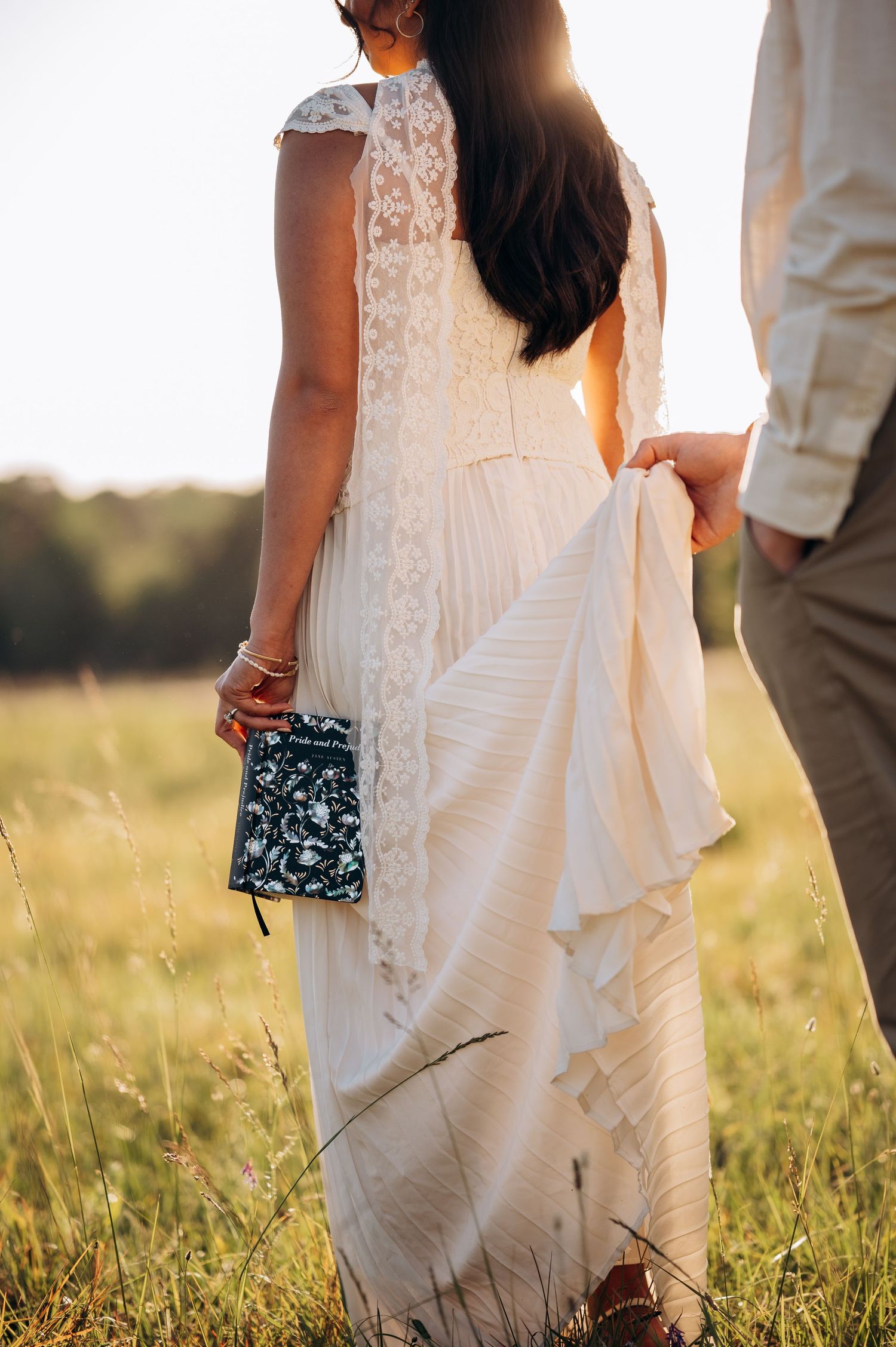 Bride in lace wedding dress holding clutch purse, walking hand in hand with groom in golden field at sunset.