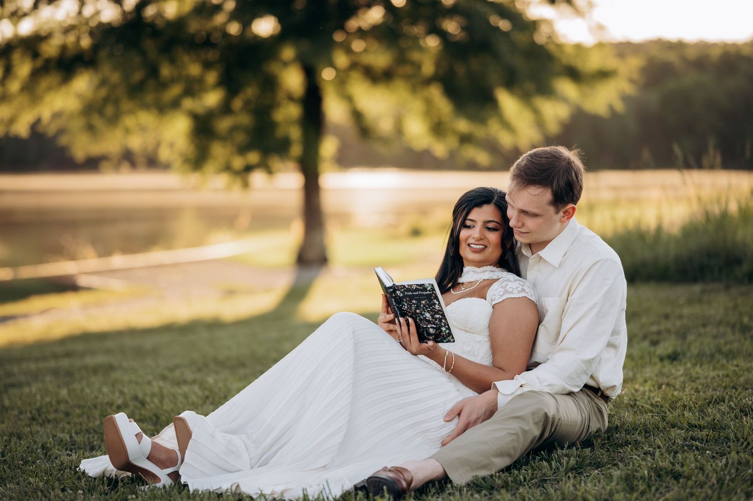 Couple in wedding attire sitting on grass by a lake at sunset, sharing a book together.