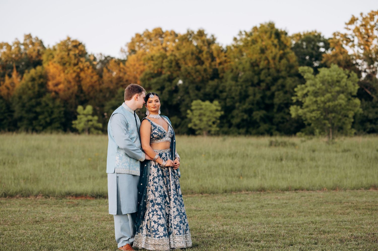 Couple in elegant Indian wedding attire embracing in a lush green field with autumn trees in background.