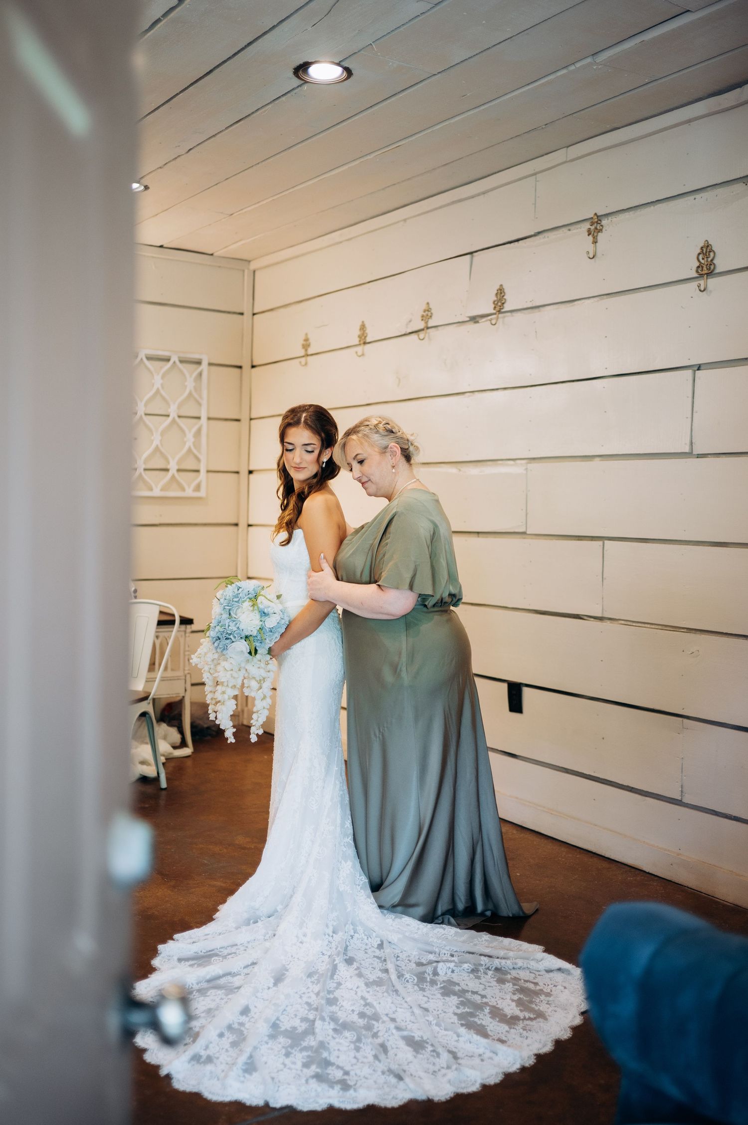 Bride in white lace gown holding blue bouquet while bridesmaid in sage green dress helps her prepare indoors.