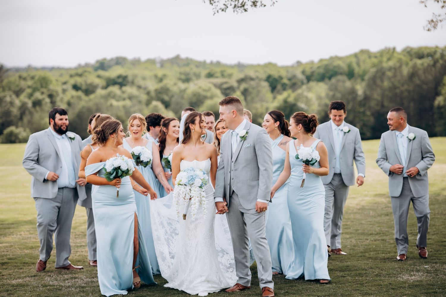 Wedding party in light blue dresses and gray suits walking outdoors on green lawn with lush trees in background.