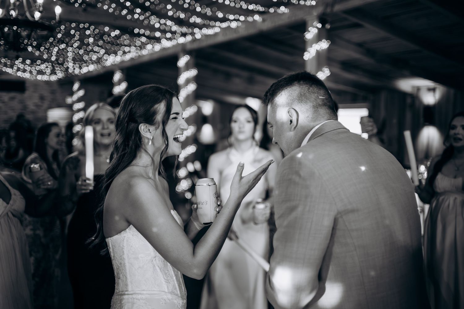 Black and white photo of bride and groom sharing a dance at wedding reception under string lights.