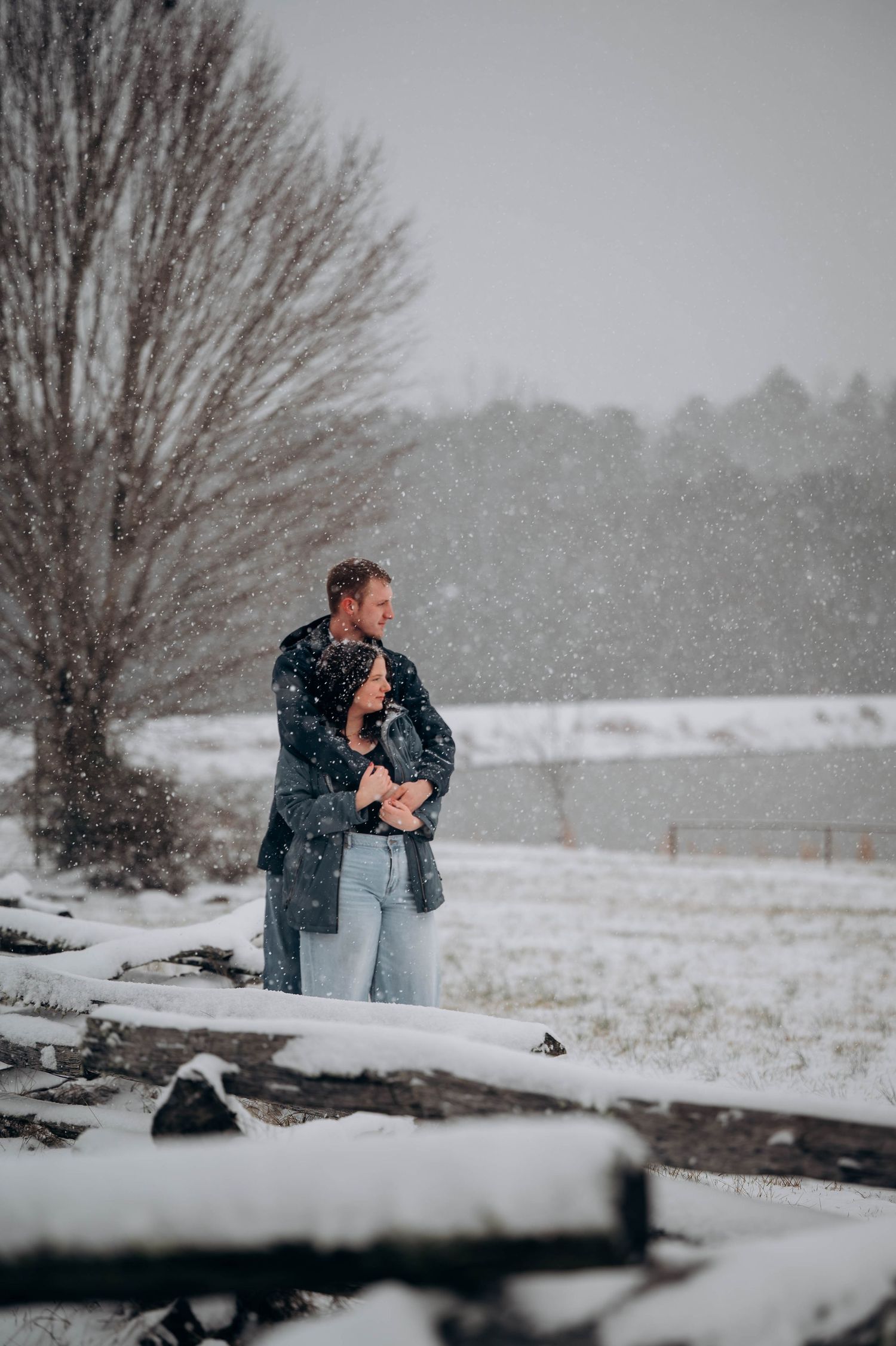 A couple stands together on a snowy hill overlooking a serene winter landscape with bare trees.