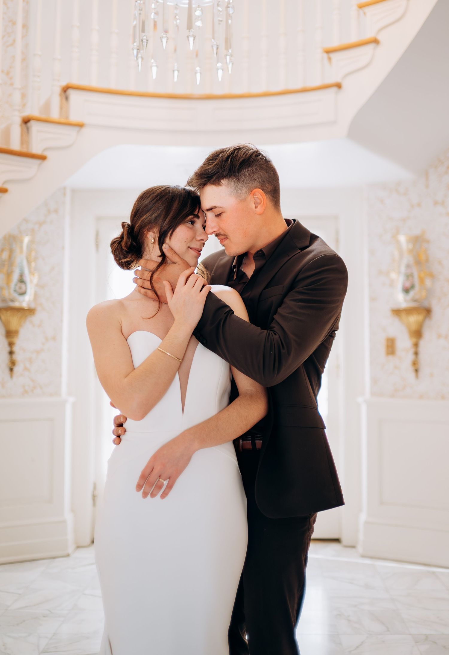Tender moment between couple in elegant white interior with crystal chandelier and curved staircase.