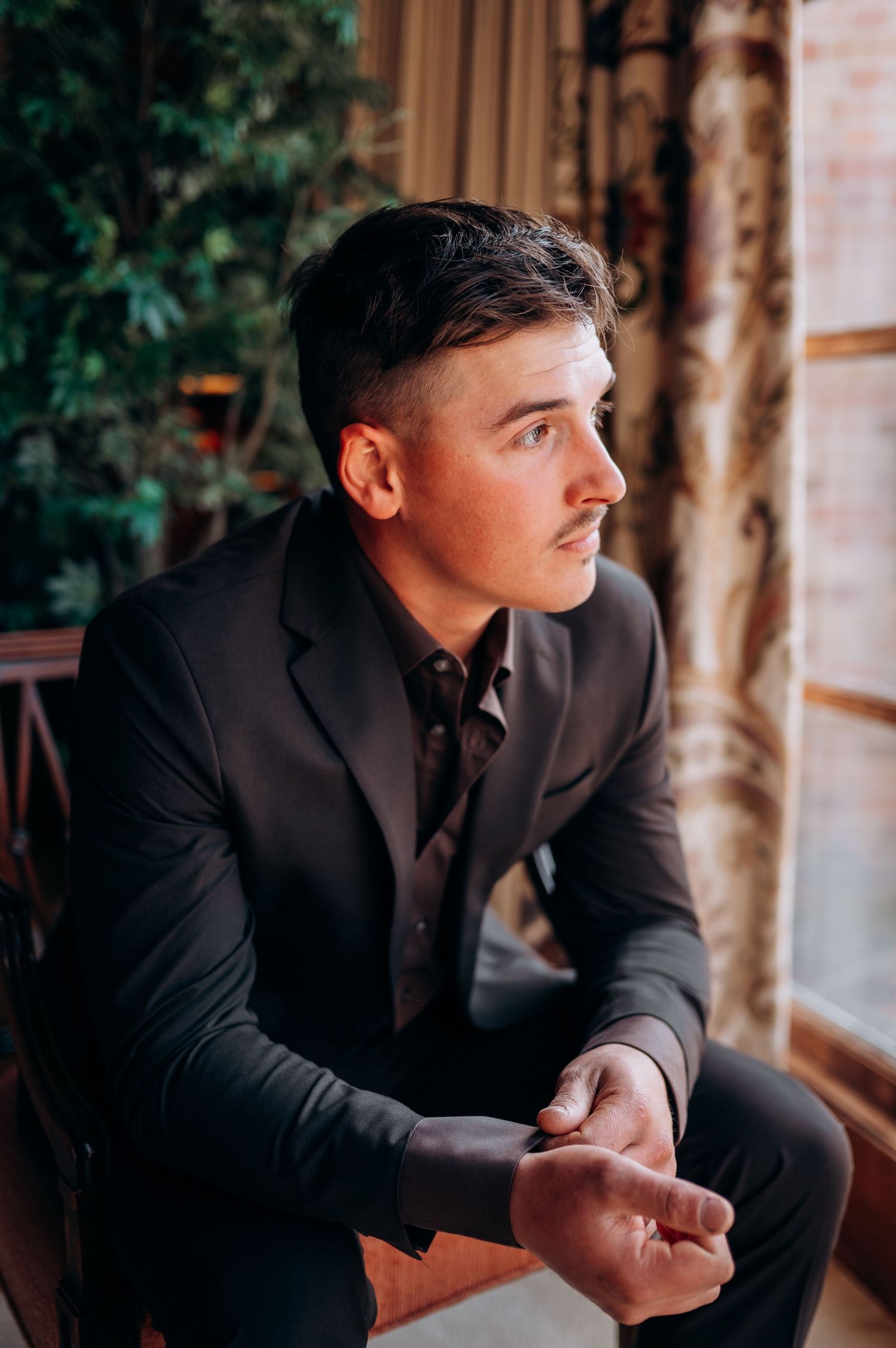 A person in a dark suit sits thoughtfully by a window with natural light streaming in.