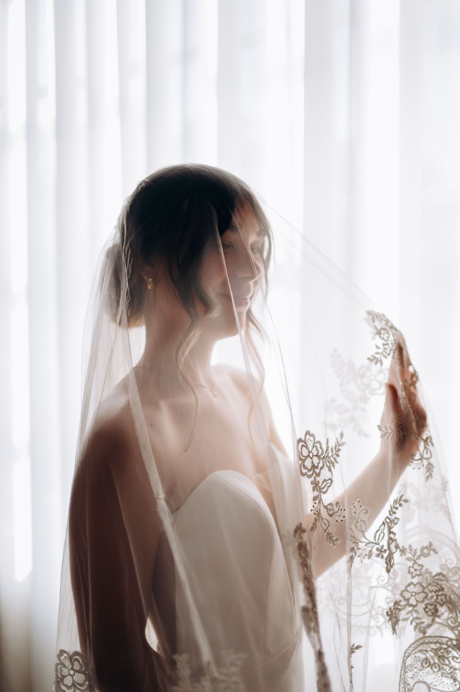 Bride in strapless gown holds lace veil near sheer curtains in soft backlit window light.