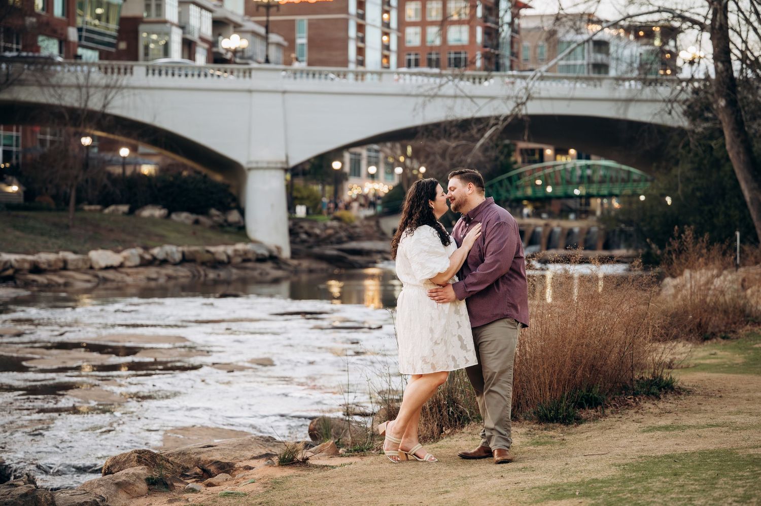 A couple shares a romantic moment under a bridge by a river during their engagement photo session.