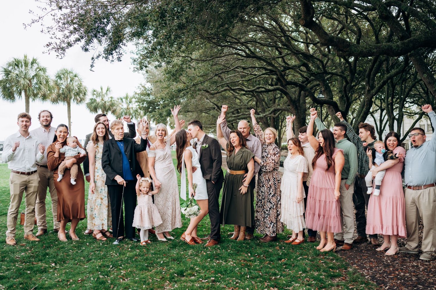 Large wedding party group photo outdoors under oak trees on a grassy lawn.