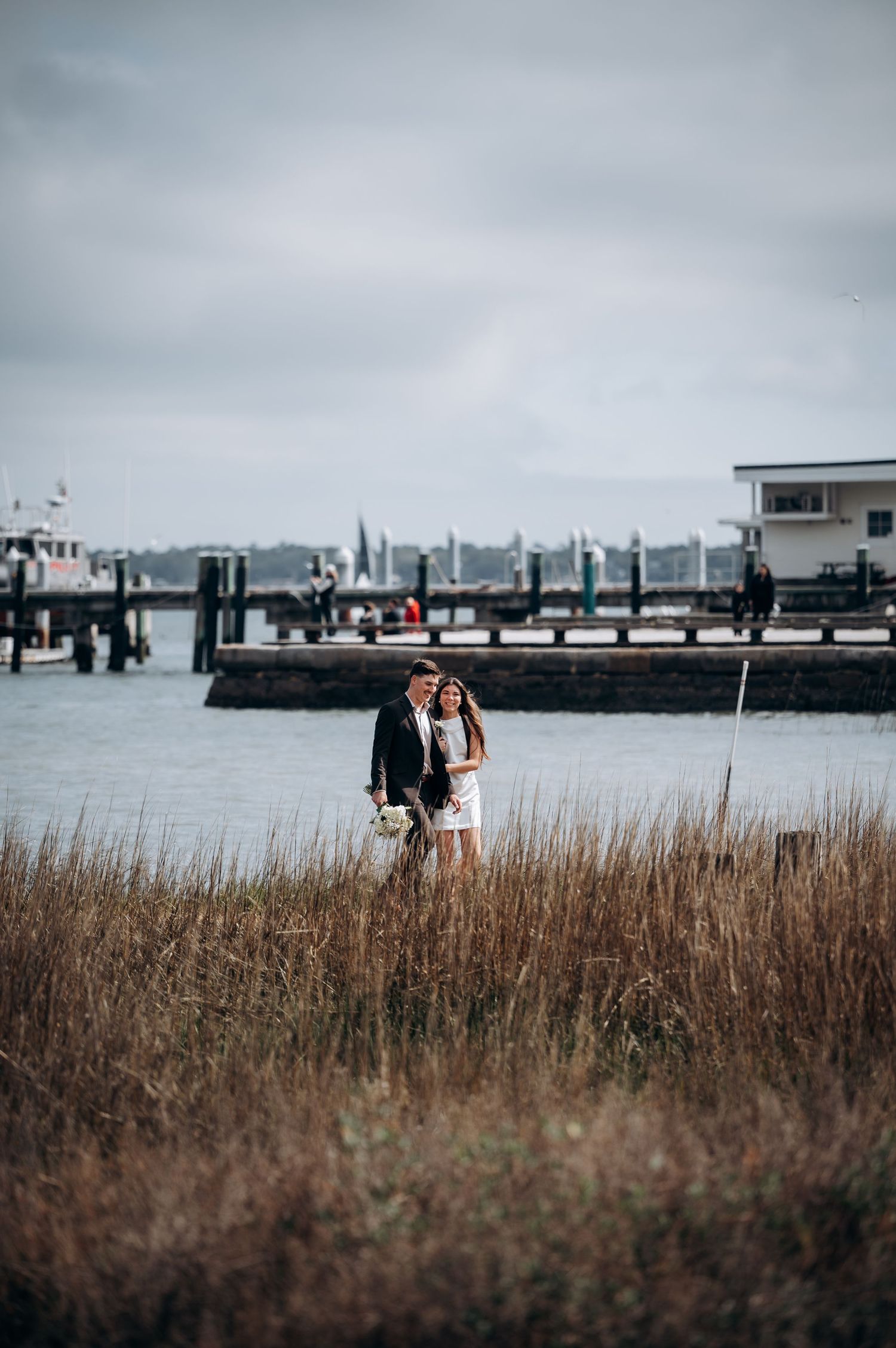 A romantic coastal scene with a couple standing among marsh grasses near a harbor.