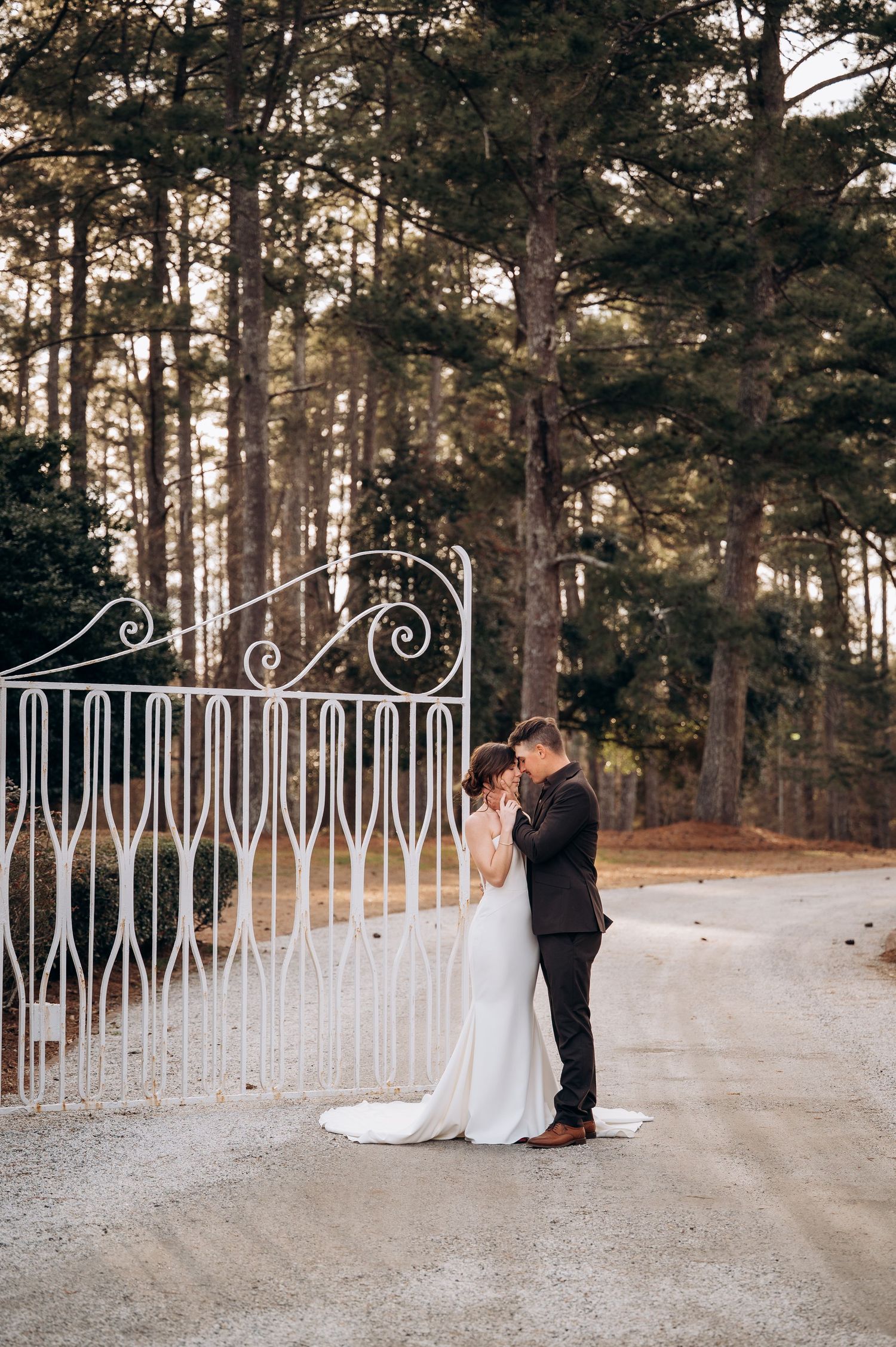Newlyweds share an intimate moment by a decorative white railing with pine trees in the background.