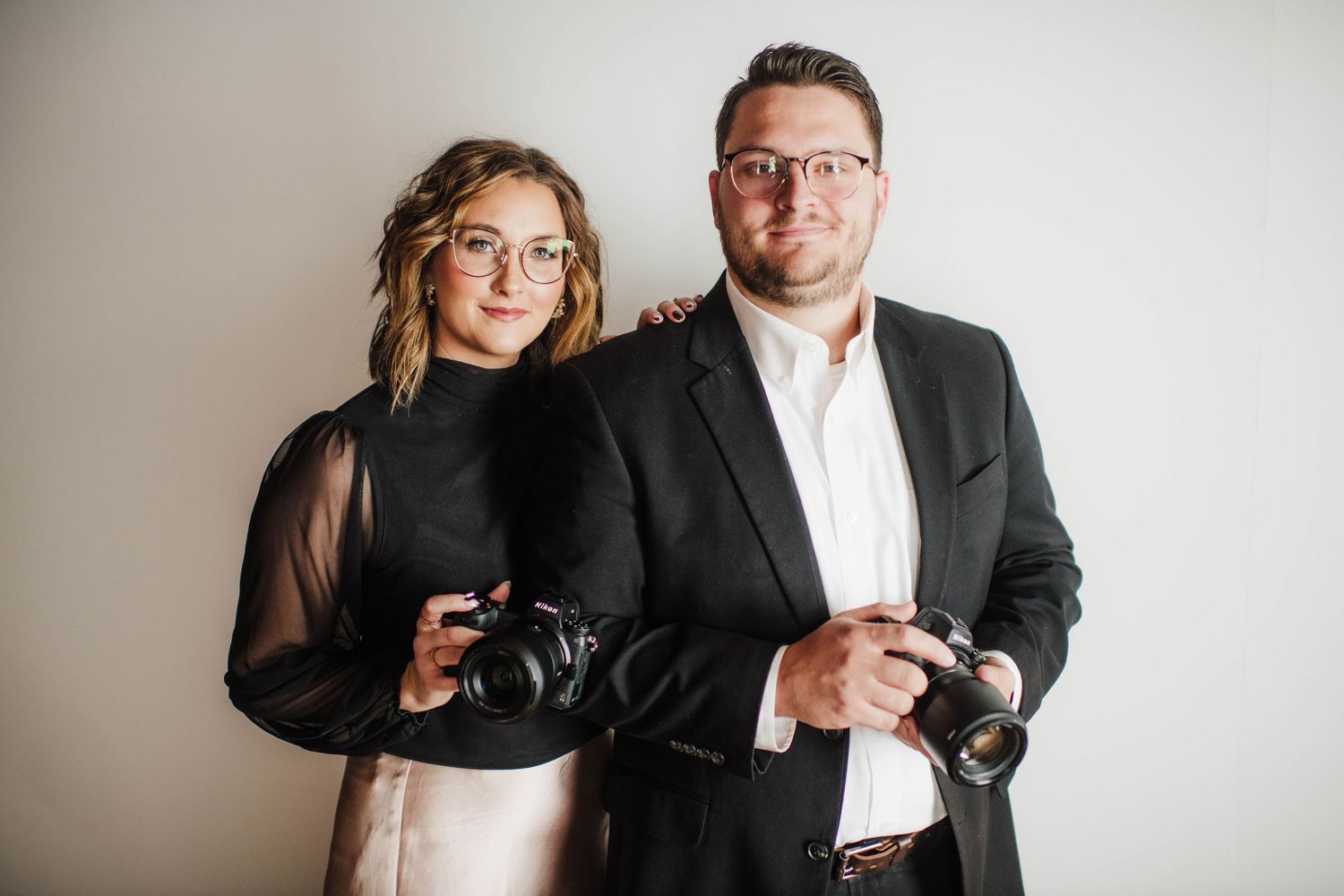 Two photographers pose with cameras in a minimalist studio setting.