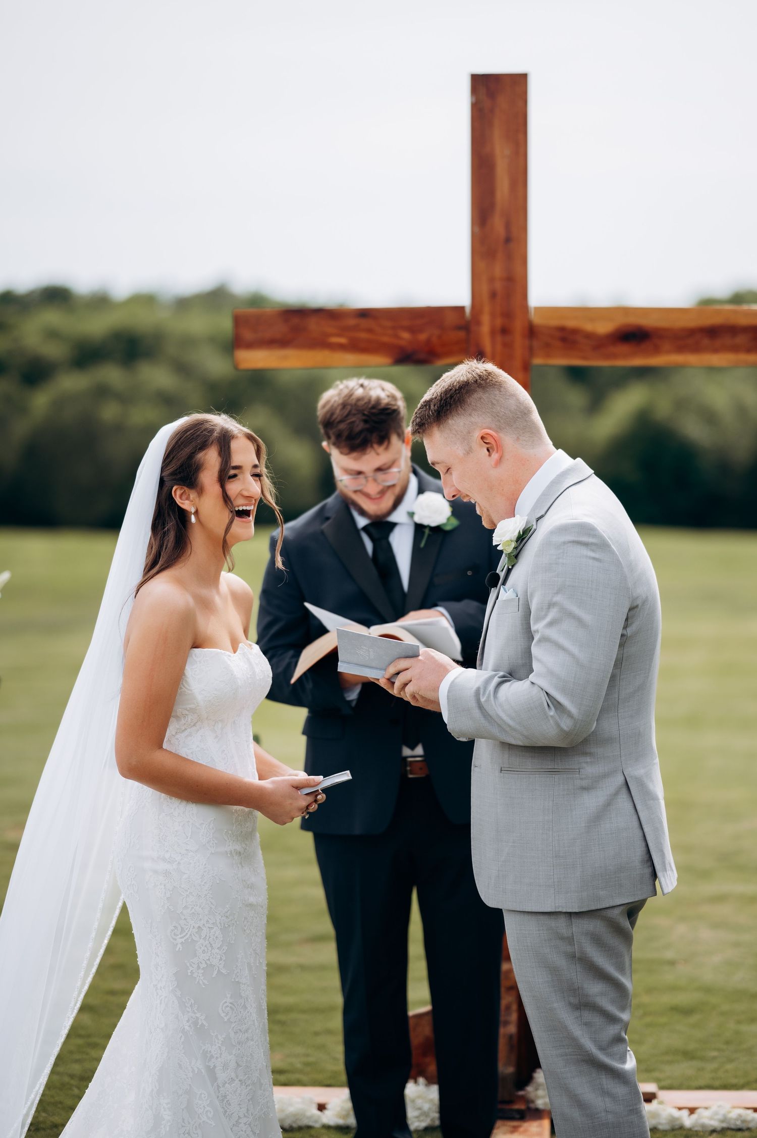 Bride and groom exchange vows before a wooden cross, officiated by a minister at an outdoor wedding.