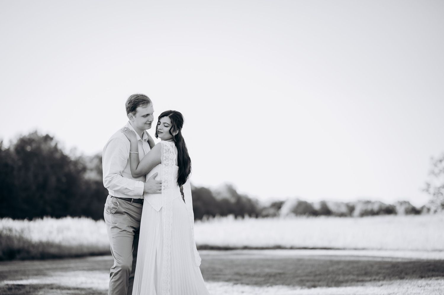 Black and white photo of a couple embracing outdoors, bride in white gown and groom in suit, near a serene lake.