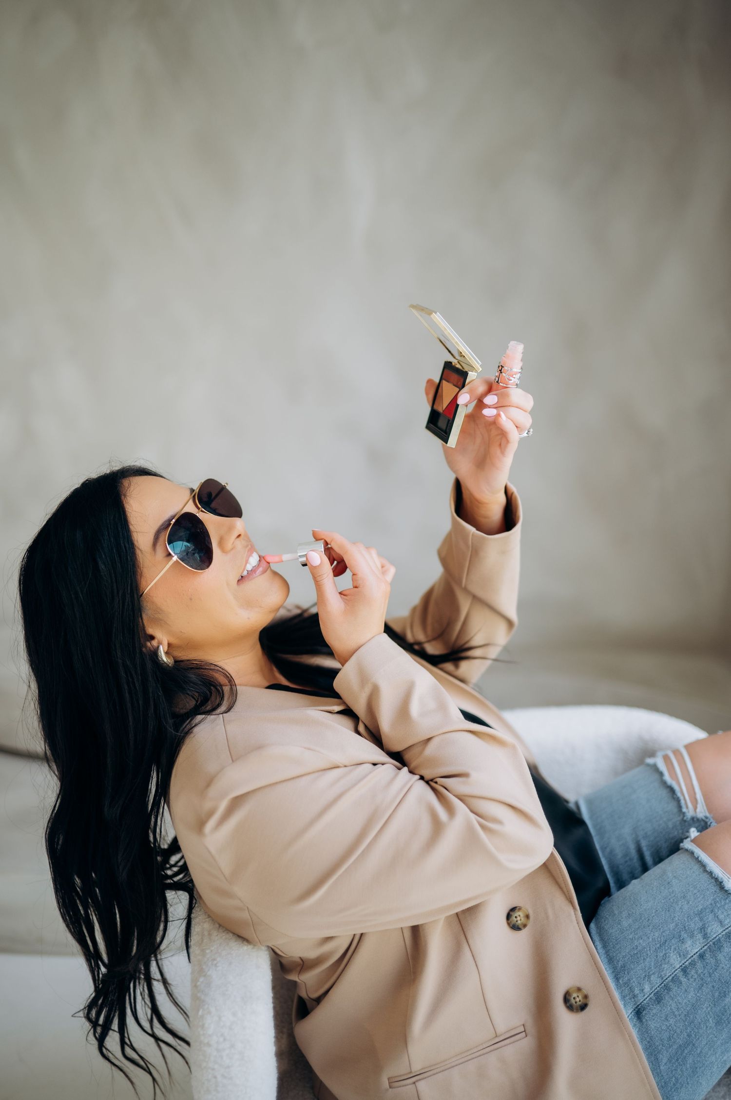 Person in beige jacket reclines playfully on couch wearing sunglasses and jeans.