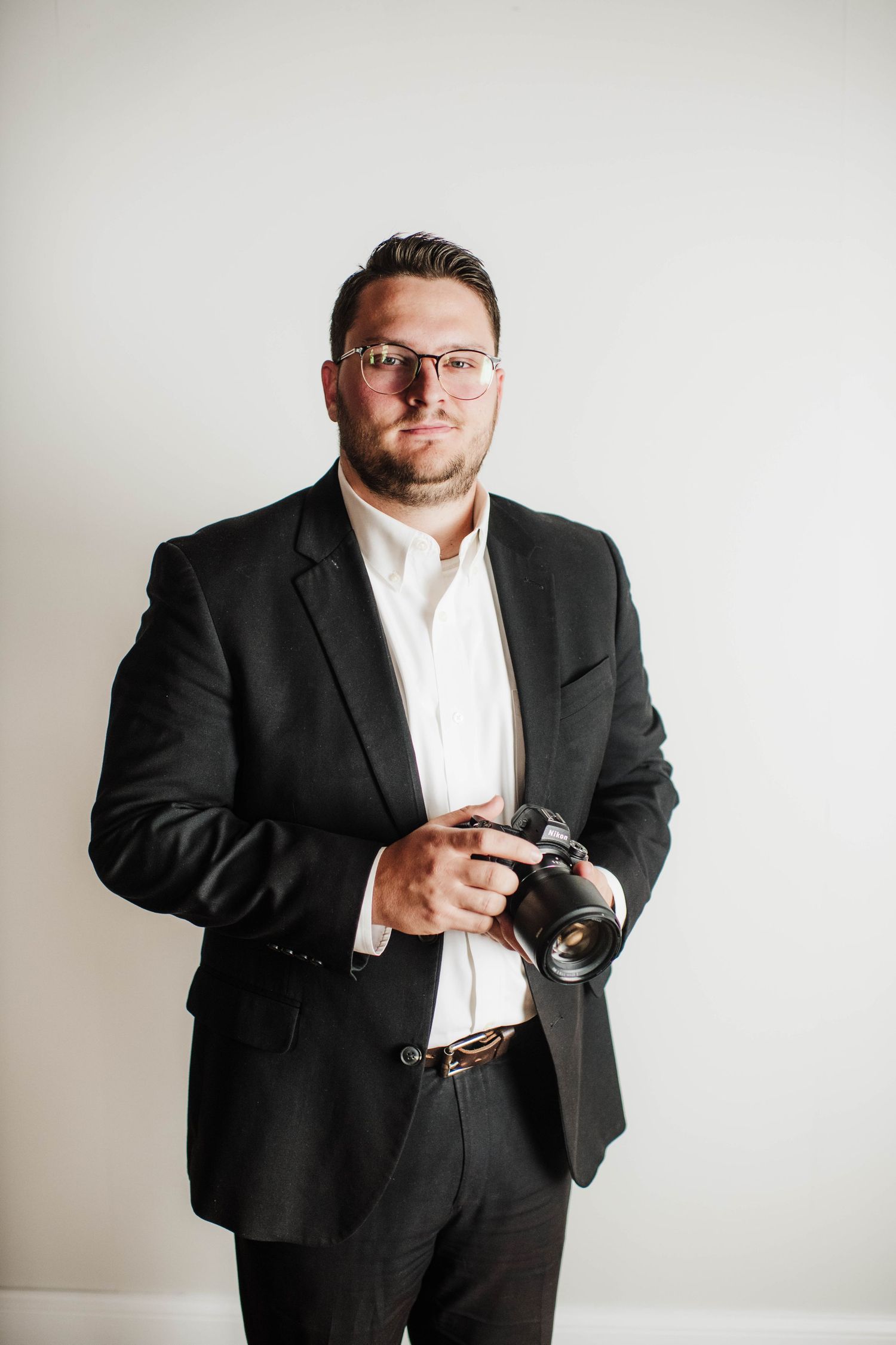 Professional male photographer in black suit and glasses holding DSLR camera, posing against white background.